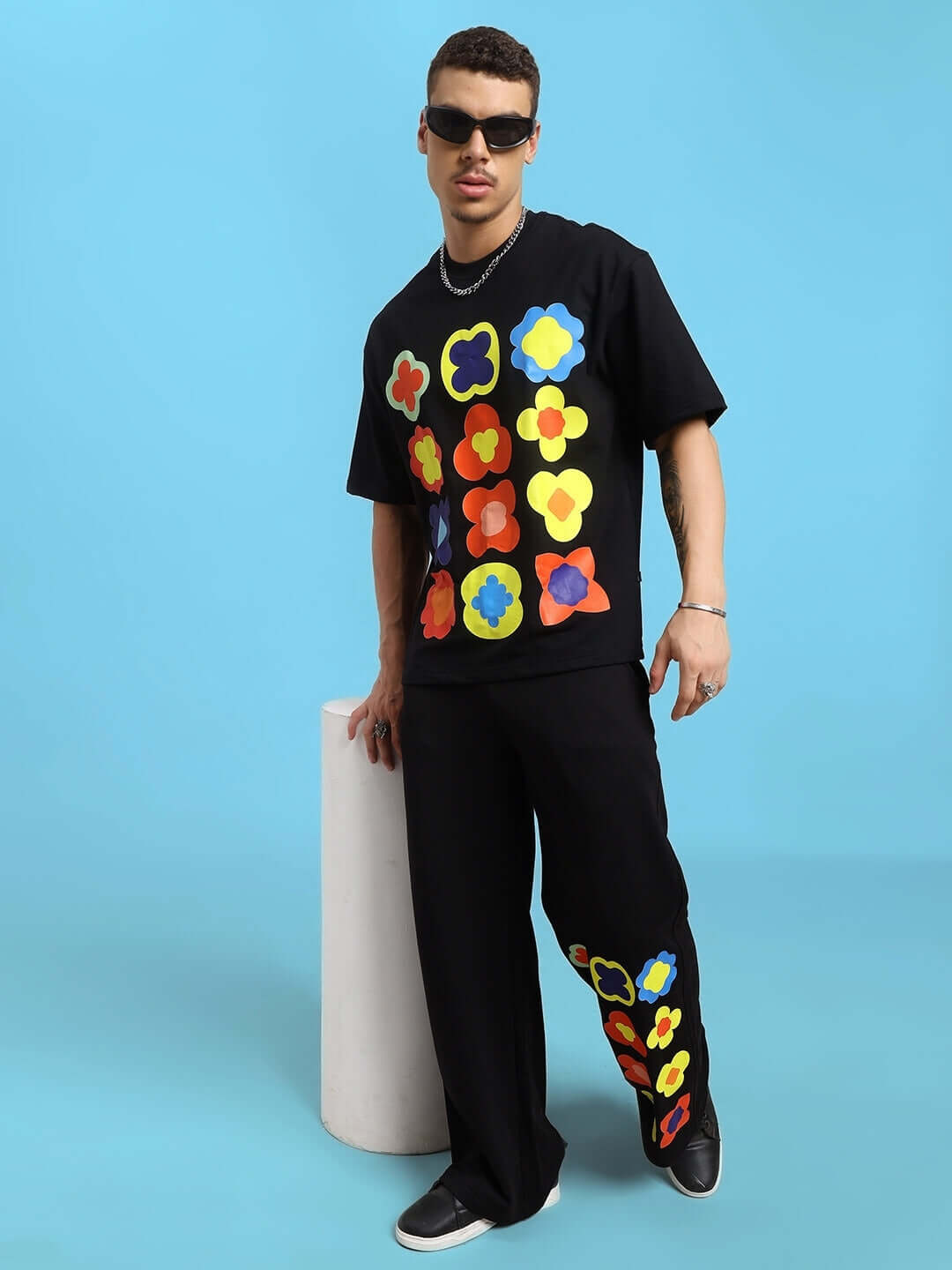 Stylish man wearing Hollister oversized black T-shirt with colorful floral patterns, paired with matching pants, against a blue background.