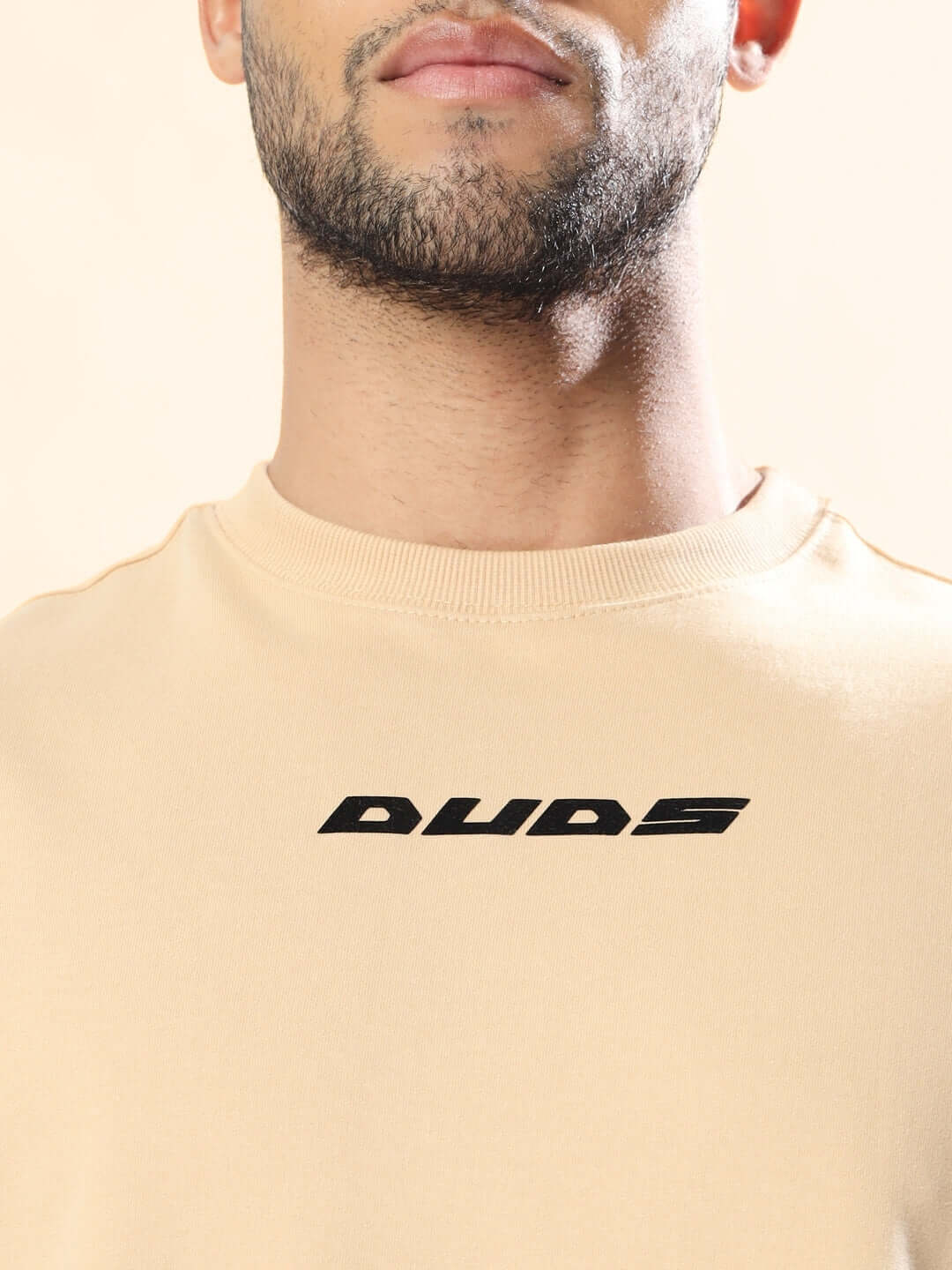 Person wearing MORE 777 OVER-SIZED T-SHIRT BEIGE made of premium heavy gauge fabric, featuring a logo on chest.