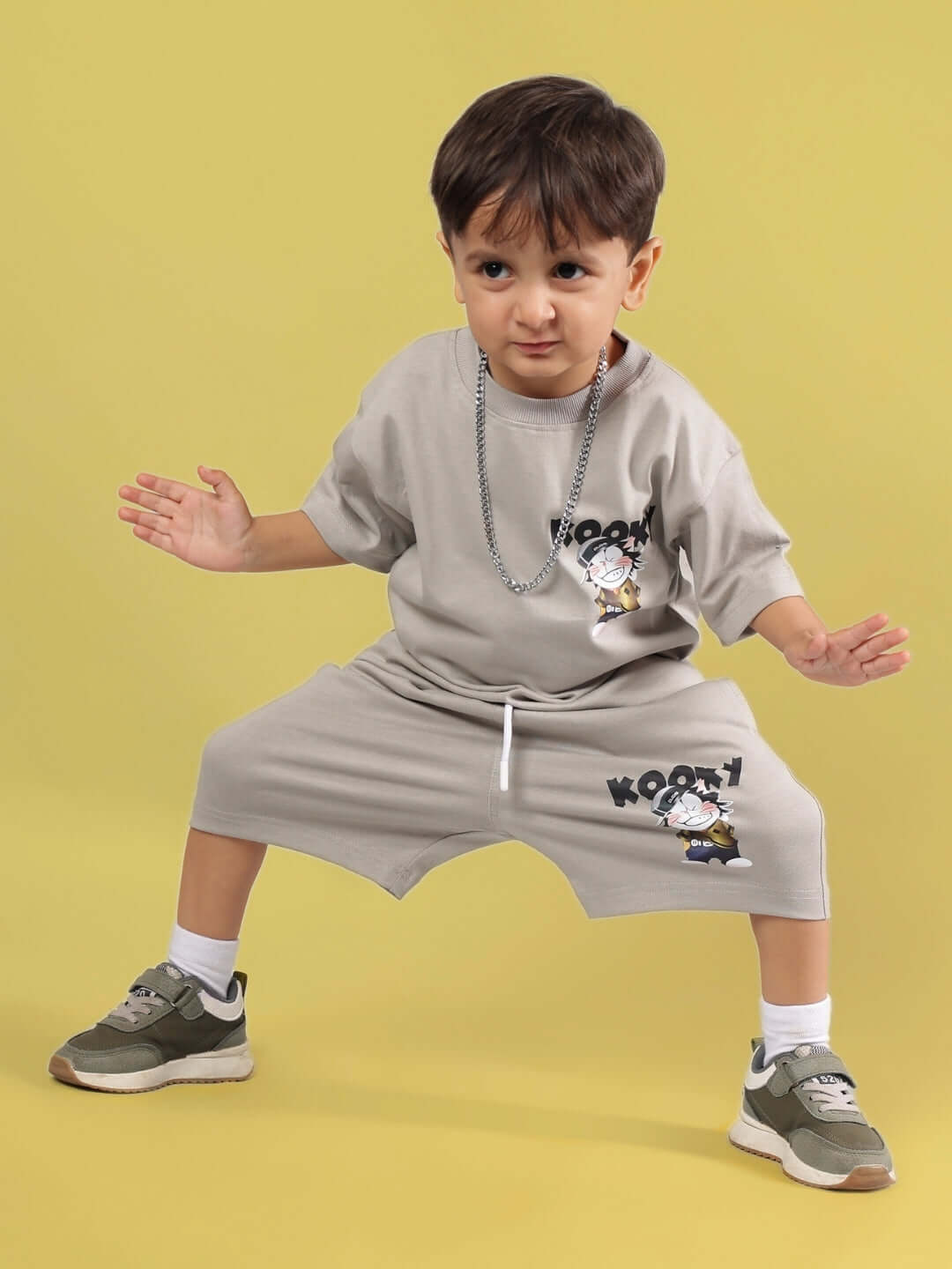 Young boy wearing a grey KOOKY T-shirt and shorts set, paired with stylish sneakers, striking a playful pose against a yellow background.