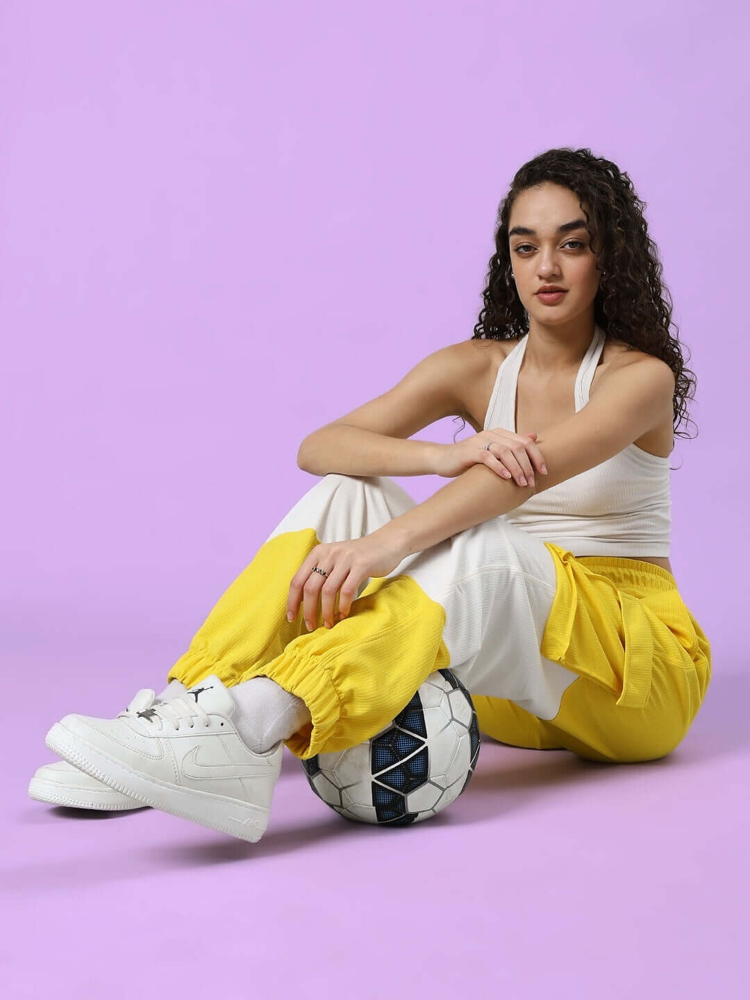 Woman wearing Women's Radiant Relaxed Fit Joggers in yellow and white, sitting with a soccer ball on a purple background.