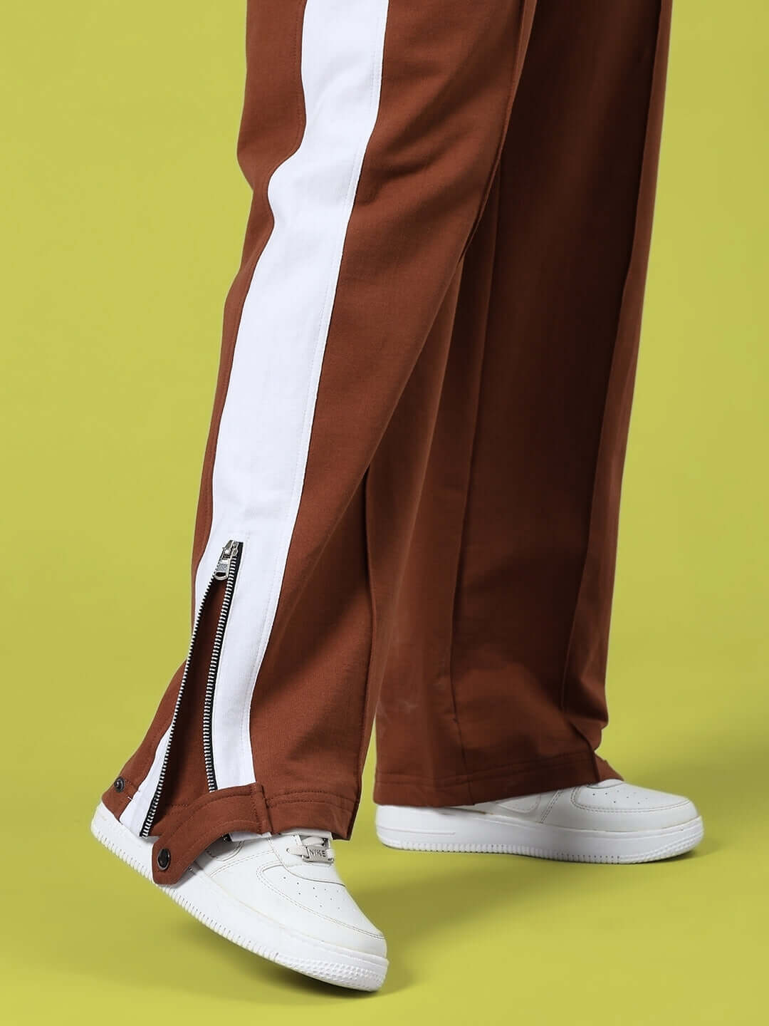 Brown women's joggers with slit zipper design, featuring white side stripes, worn with white sneakers on lime green background.