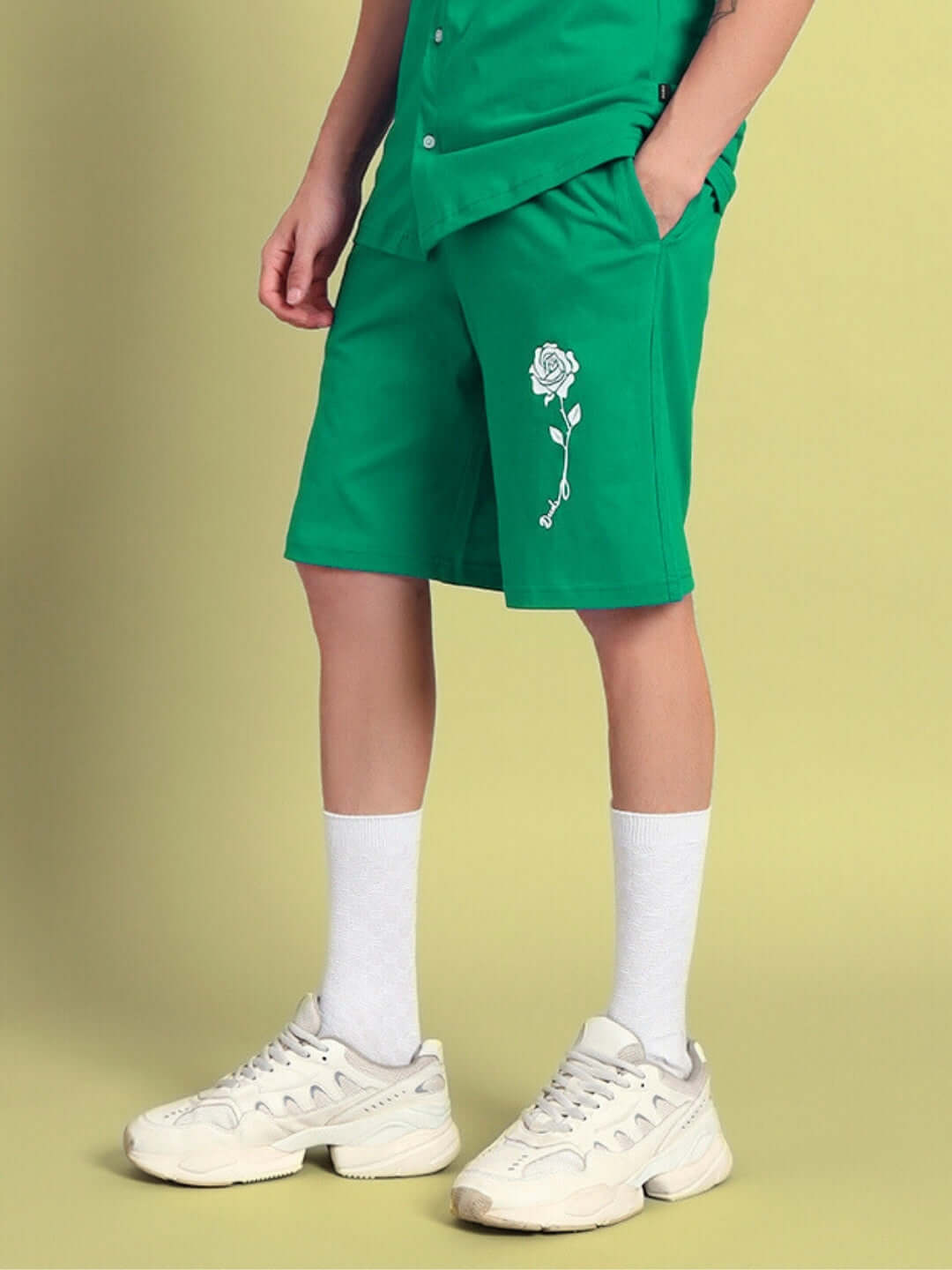 Person wearing green Limbo Summer Shorts with white embroidered design, styled with white socks and sneakers on light backdrop.