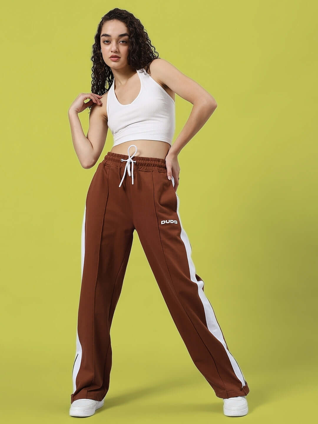 Woman wearing brown women's wonder slit zipper joggers with white stripes by DUDS, in fashionable pose against green background.
