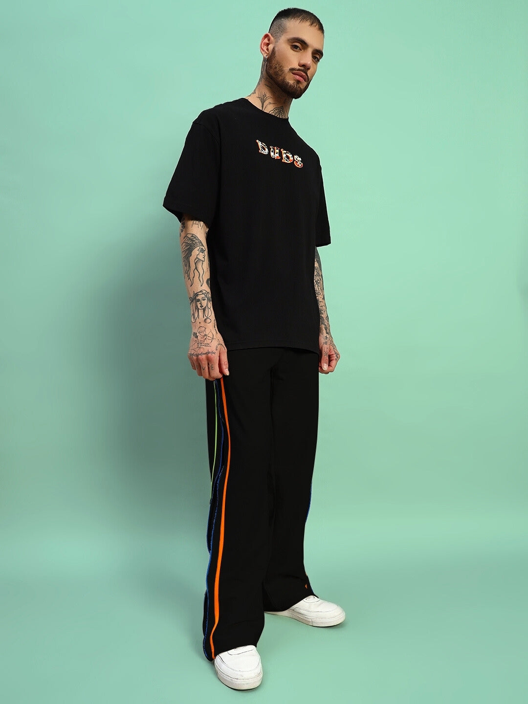 Man wearing black IMPERIAL SIDE ZIPPER JOGGERS by DUDS, showcasing side stripe design and relaxed fit, standing against mint green backdrop.