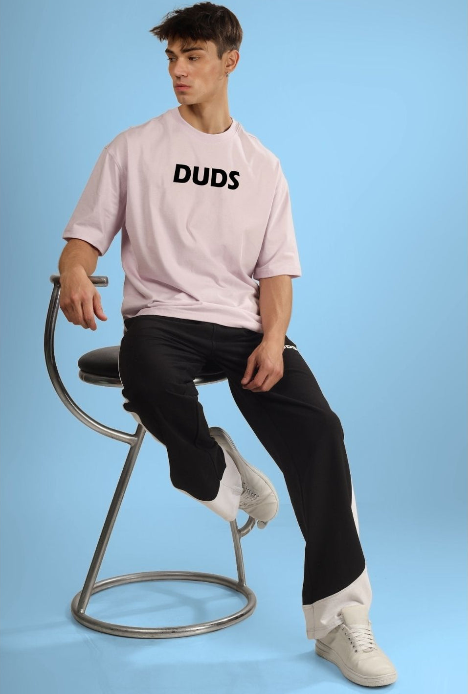 Model wearing casual pink t-shirt with "DUDS" print, styled with black pants and white sneakers, seated on a modern metal chair.