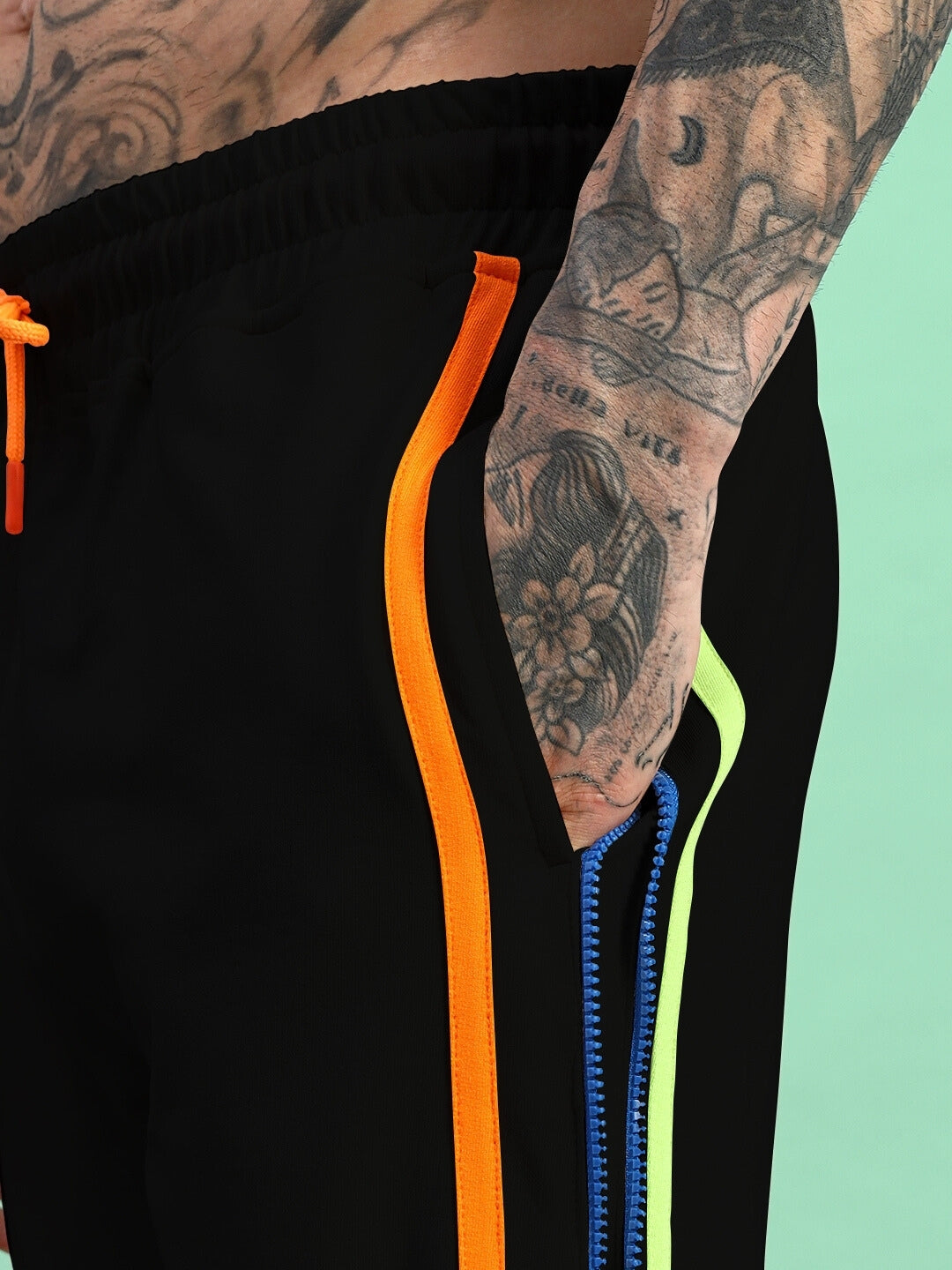 Close-up of black joggers with side zipper detail, featuring vibrant orange and neon accents on a tattooed model. Made to order by DUDS.