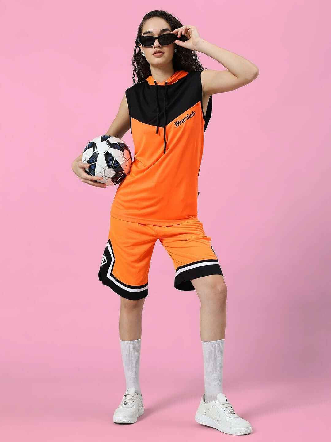 Model wearing Women's Armado Regular Fit Shorts in orange and black, holding a soccer ball against a pink background.