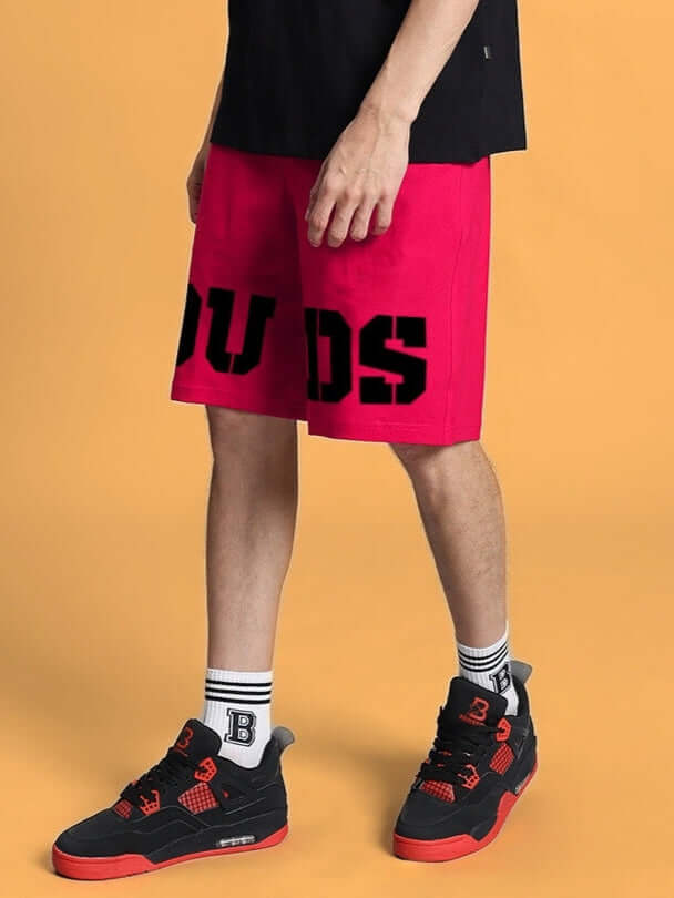 Model wearing pink Cherry Blossom Regular Fit Shorts with bold DUDS logo, paired with black and red sneakers against a yellow background.