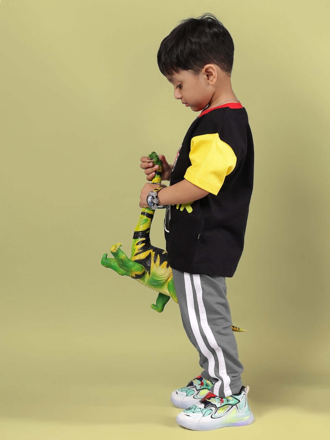 Child wearing black and grey co-ord set holding green dinosaur toy against a yellow background.