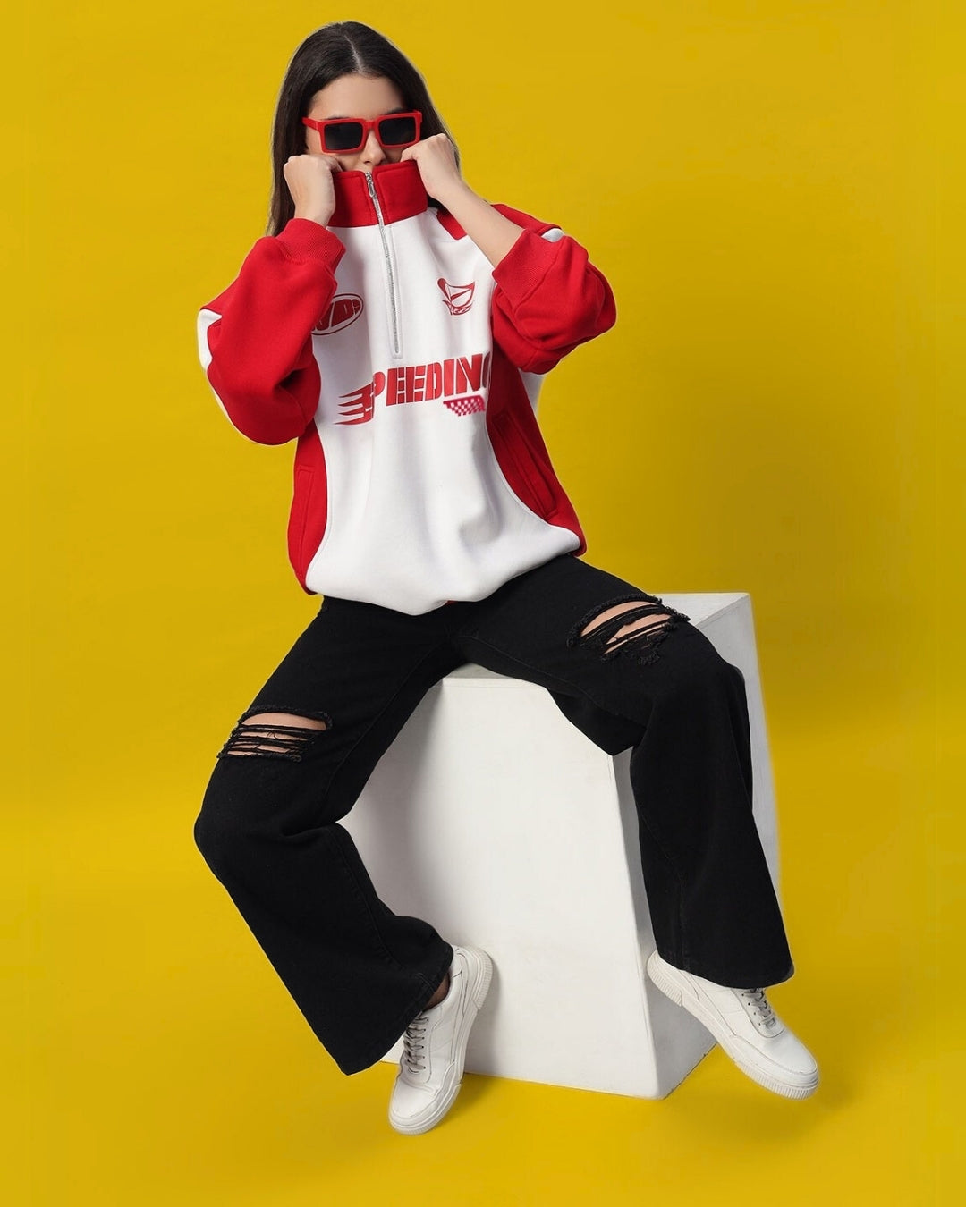 Woman wearing red and white colorblock jacket and black jeans, sitting stylishly on white cube with a vibrant yellow background.