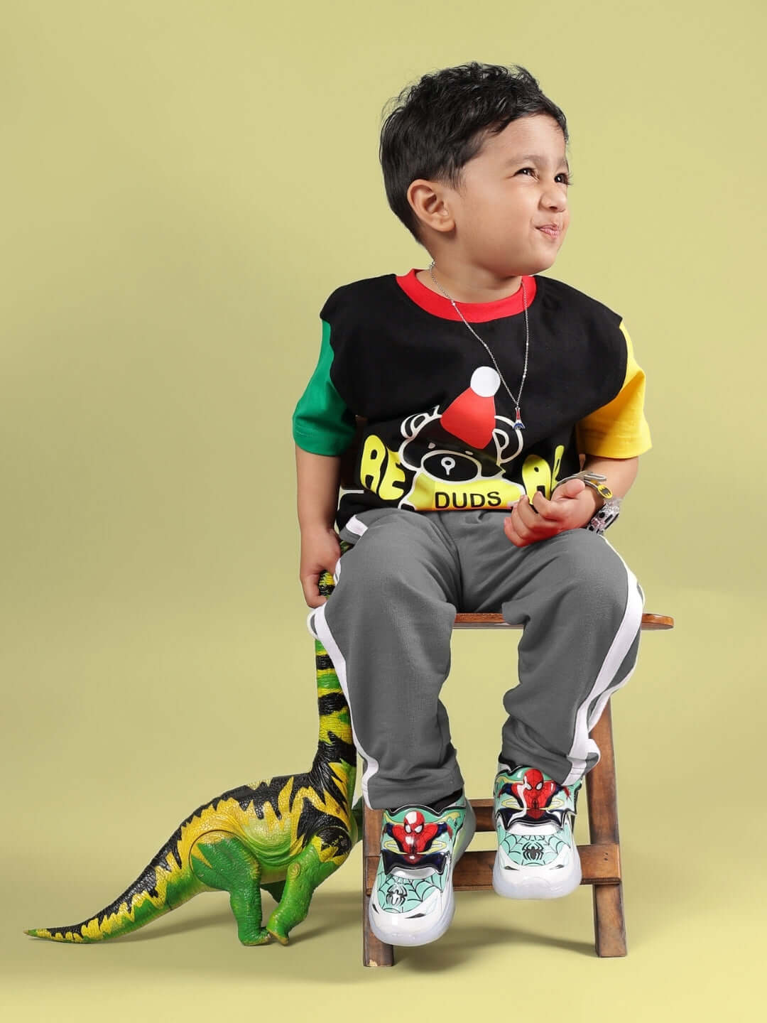 Young child wearing a colorful black and grey MOCHA CO-ORD outfit with dinosaur toy, sitting on a stool against a yellow background.