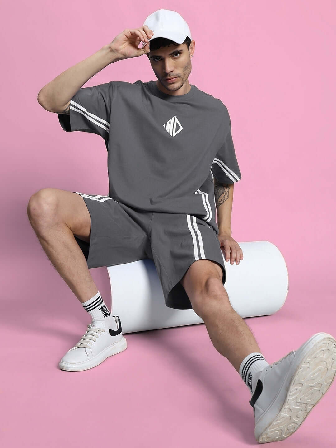 Model wearing DUDS Outlook Regular Fit Grey Shorts, styled with white sneakers, sitting against a pink backdrop.