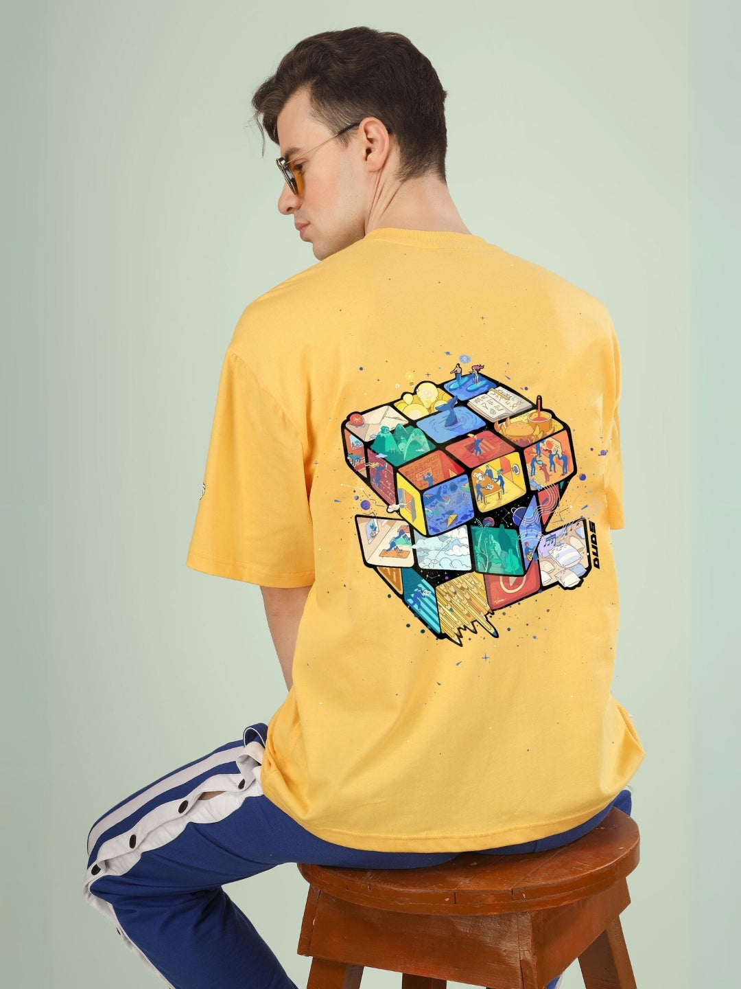 Person wearing RUBIK REVOLUTION OVERSIZED T-SHIRT YELLOW with artistic cube design, sitting on stool.