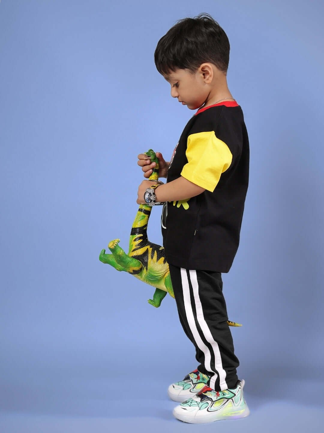 Child wearing black MOCHA CO-ORD outfit with yellow sleeves, holding a green dinosaur toy, standing against a blue background.