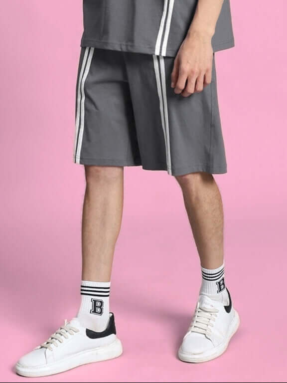 Model wearing Outlook regular fit grey shorts, featuring white striped design, paired with white sneakers and socks on pink background.