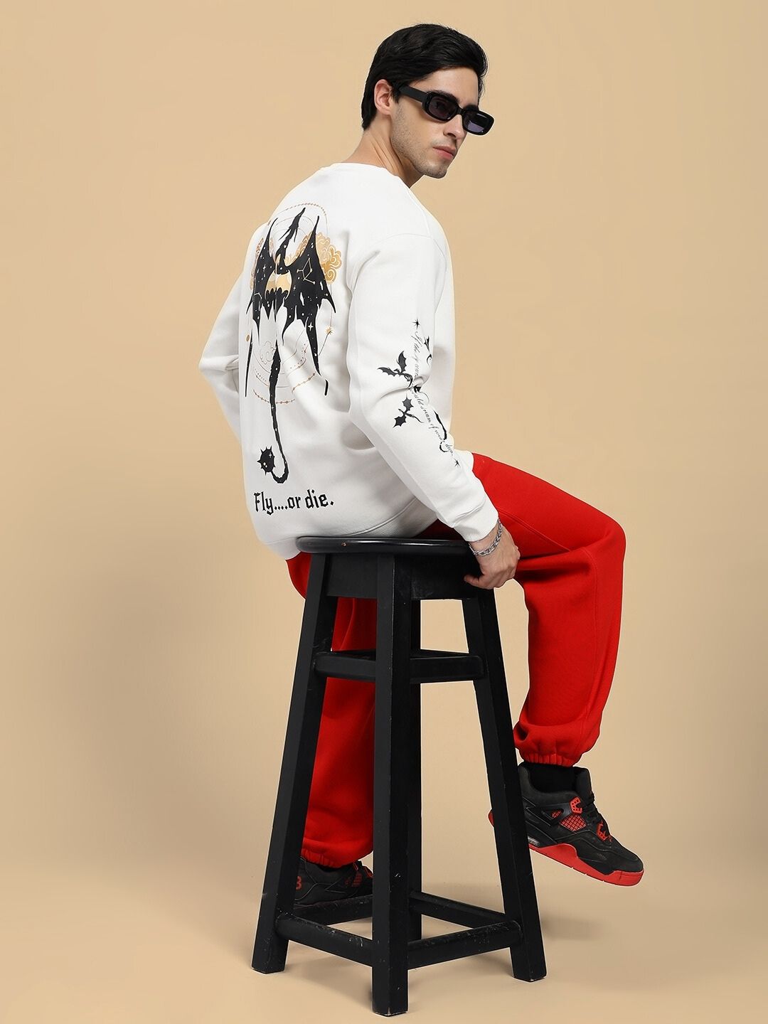 Person wearing a white sweatshirt with black graphics and red pants sitting on a black stool against a beige background.