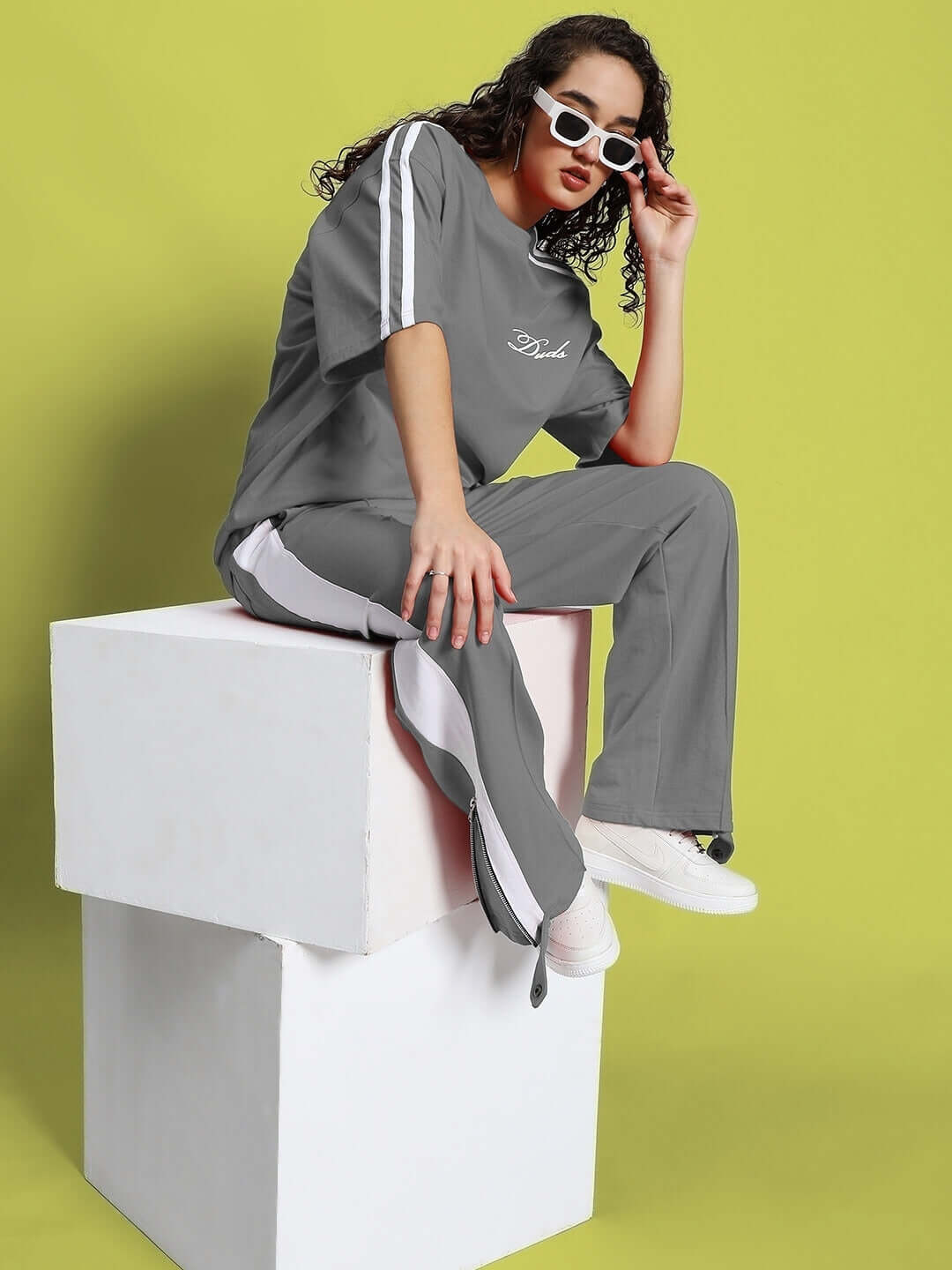 Woman in grey-white slit zipper joggers and top, sitting stylishly on a platform against a vibrant background, wearing sunglasses.
