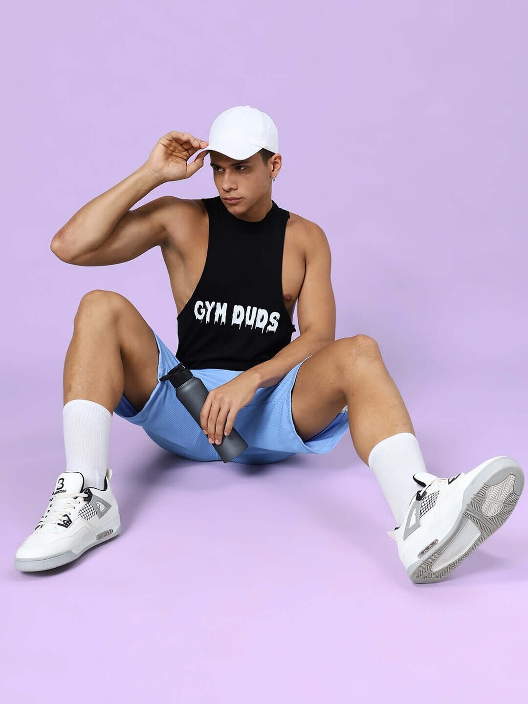 Man wearing Crunch Gym Co-Ord Set in black and blue, sitting on pastel background with cap and sneakers, holding water bottle, fitness apparel.