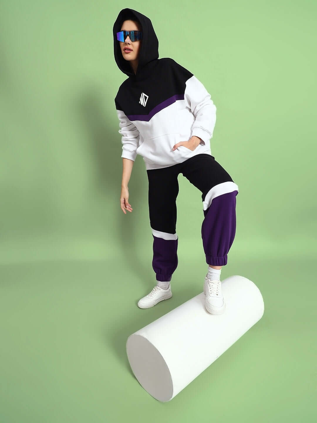 Model wearing women's Harley fleece colorblock jogger in purple, standing on cylinder, showcasing stylish streetwear fashion against green background.