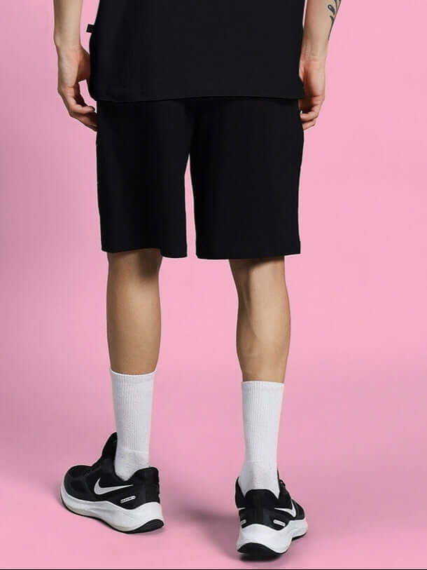Person wearing black summer shorts from Squid Game collection, standing against a pink background, showcasing casual sporty style.