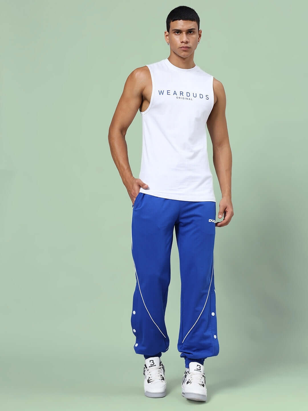 Model wearing Kickshaw relaxed fit white tank top and blue joggers set with sneakers, showcasing comfortable, unisex apparel.