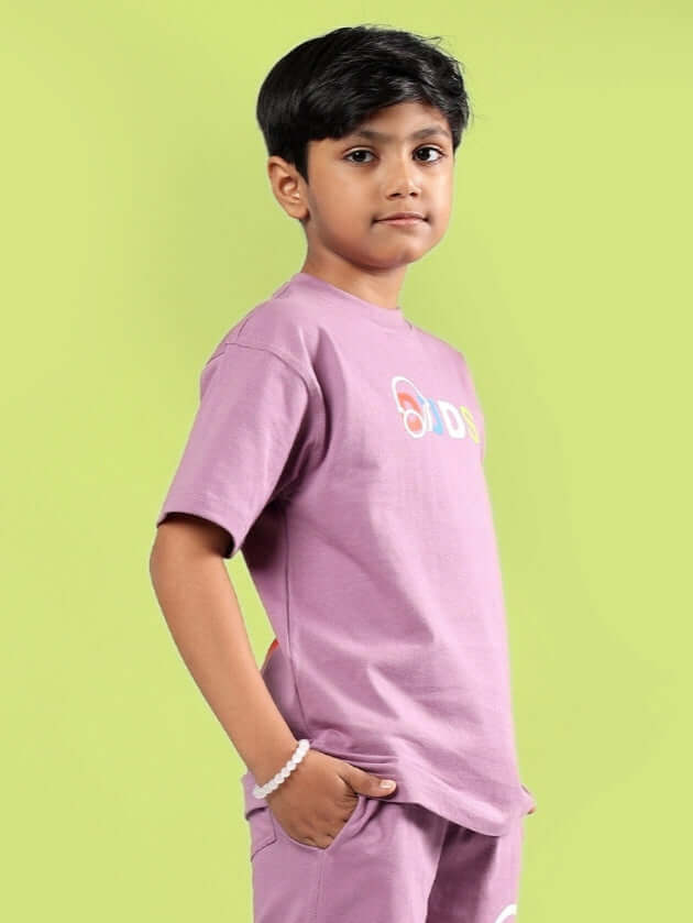 Young boy wearing STOIC purple t-shirt from DUDS, standing against a green background.