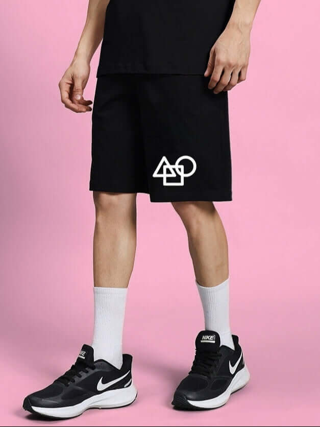 Man wearing black "SQUID GAME SUMMER SHORTS" with white symbols, standing against pink background, showcasing customizable fit options.