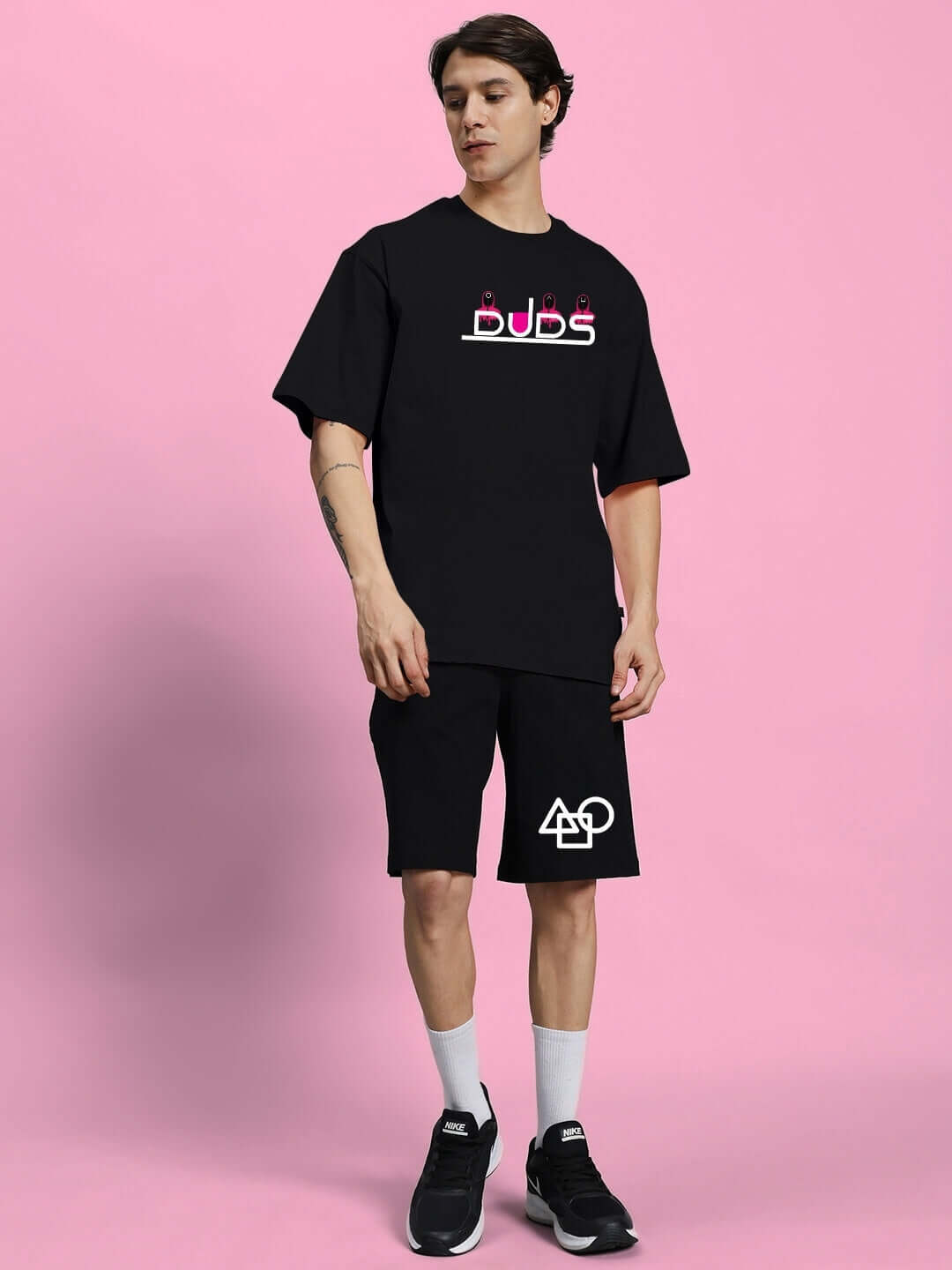 Man wearing Squid Game summer co-ord set in black with graphic prints, standing against a pink background, showcasing casual style.