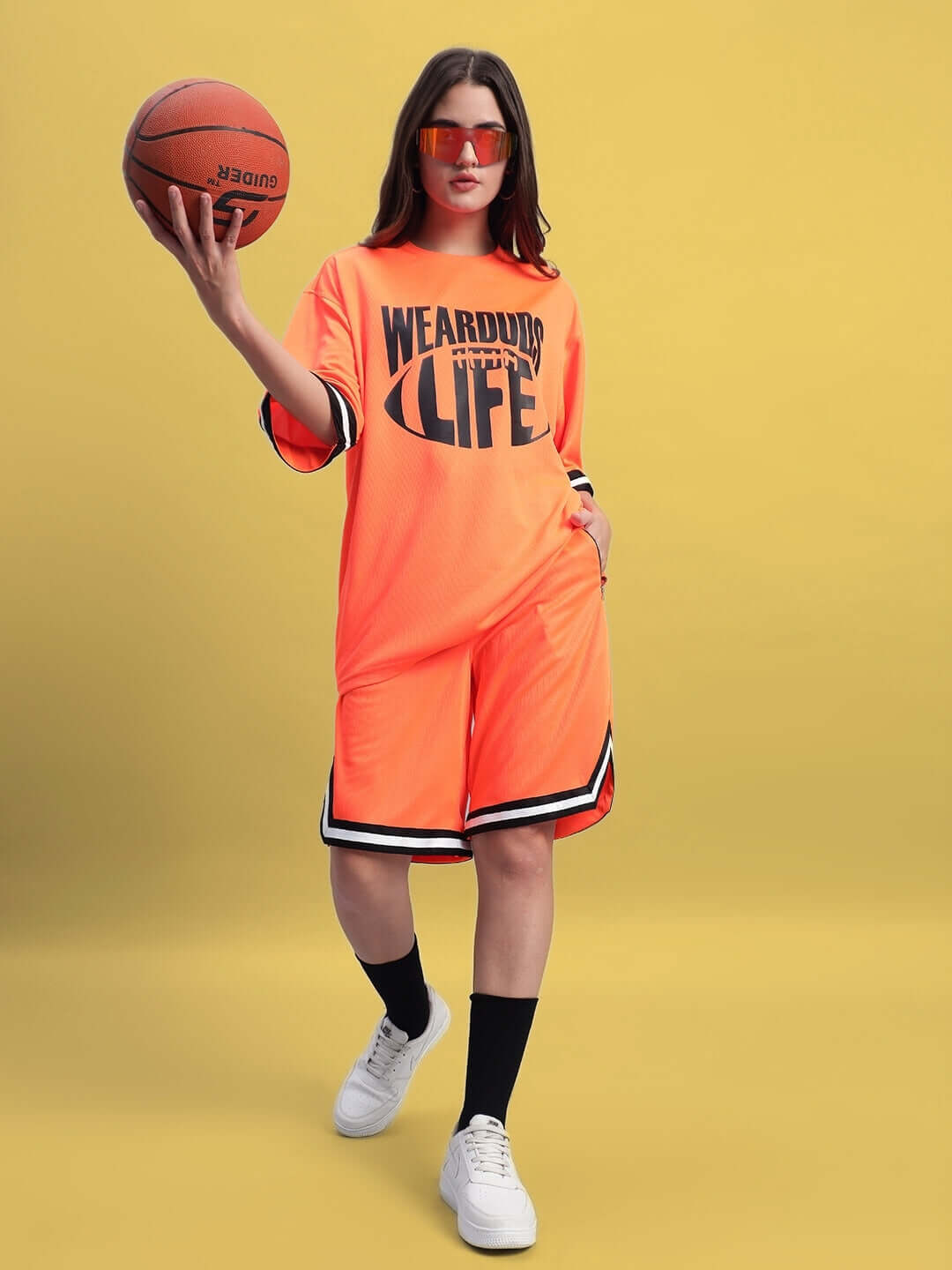 Woman wearing orange regular fit shorts holding basketball, standing against yellow background.