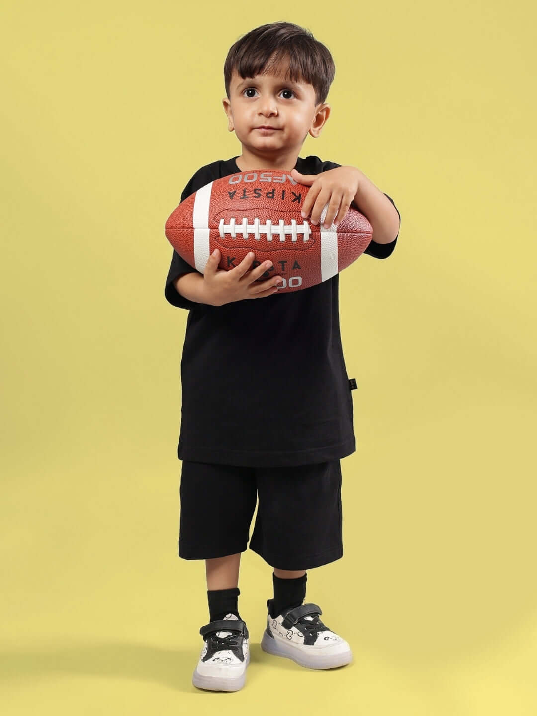 Young child wearing a black t-shirt and shorts playing with a football, standing against a yellow background.