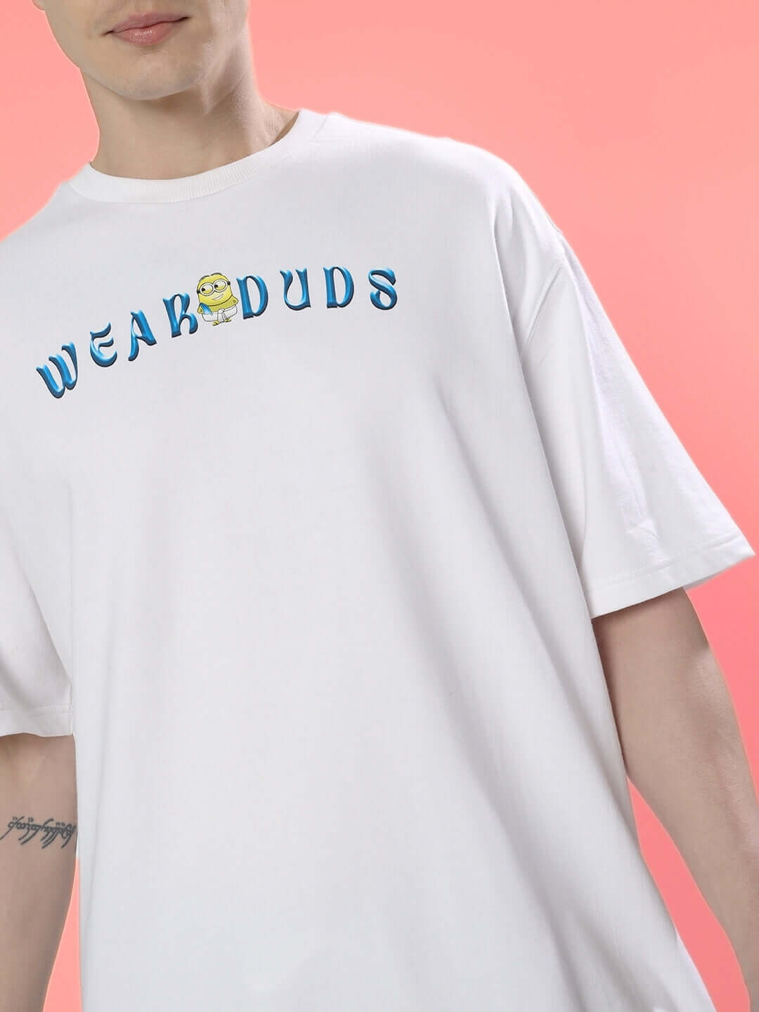 Person wearing SUBRAMIN MINION OVER-SIZED T-SHIRT WHITE with "WEAKDUDS" graphic, against pink background.