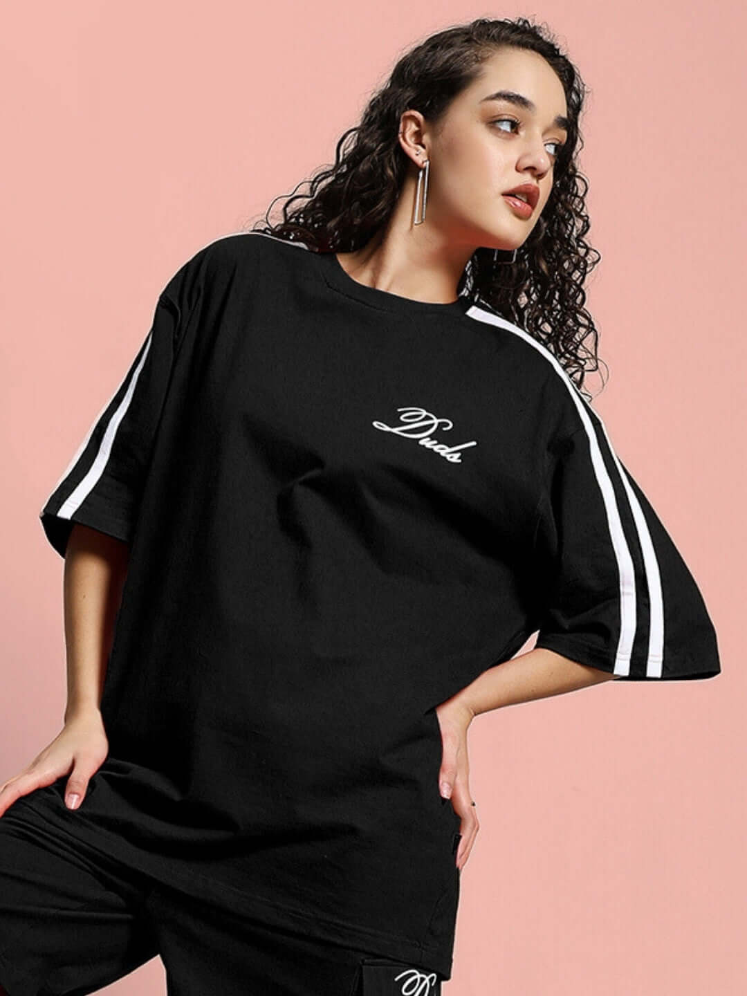 Black oversized women's t-shirt, premium cotton blend, with white stripe details, styled on a model against a pink background.