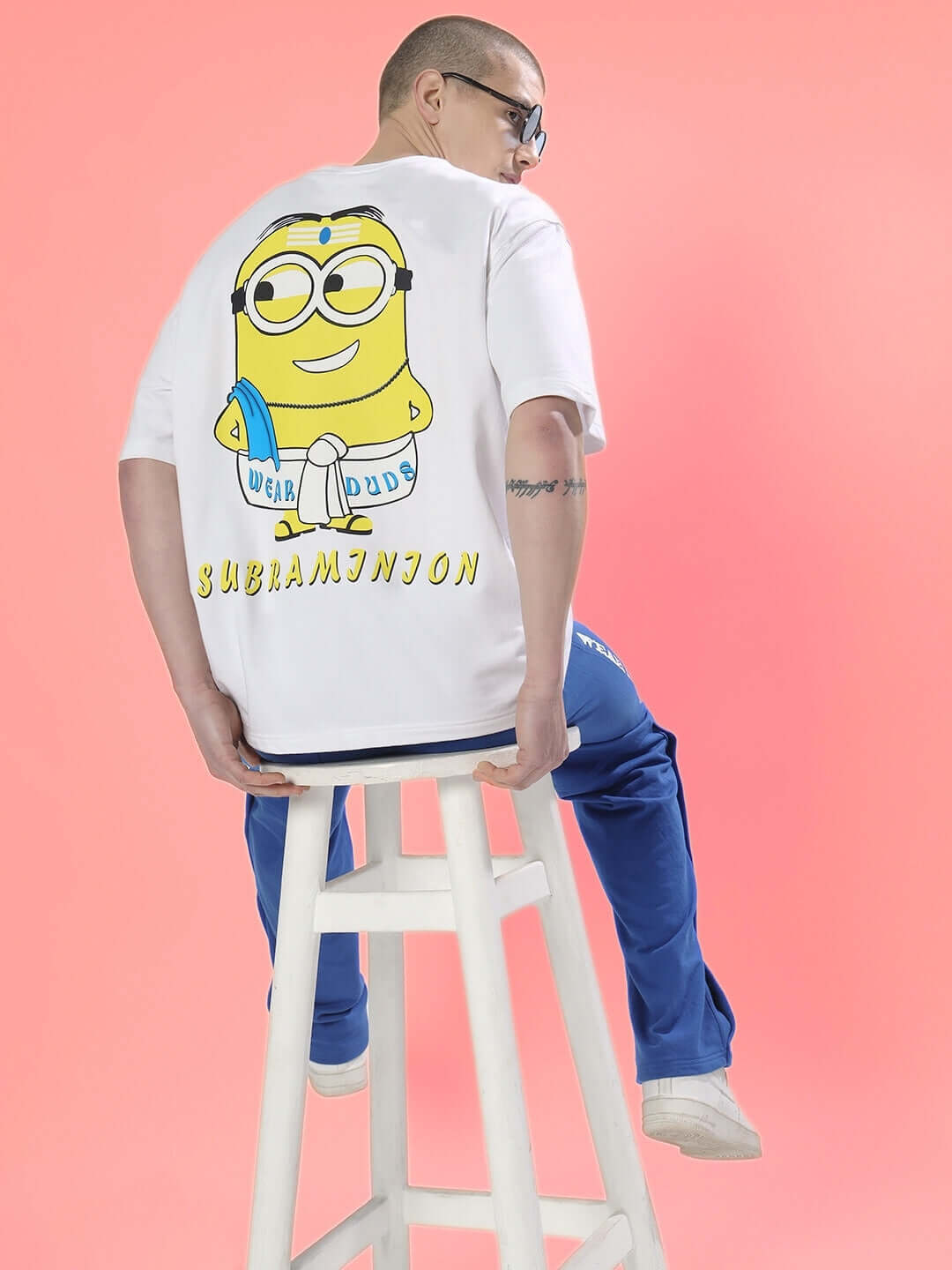 SUBRAMIN MINION OVER-SIZED T-SHIRT WHITE with graphic design on back, worn by a person sitting on a stool against a pink background.