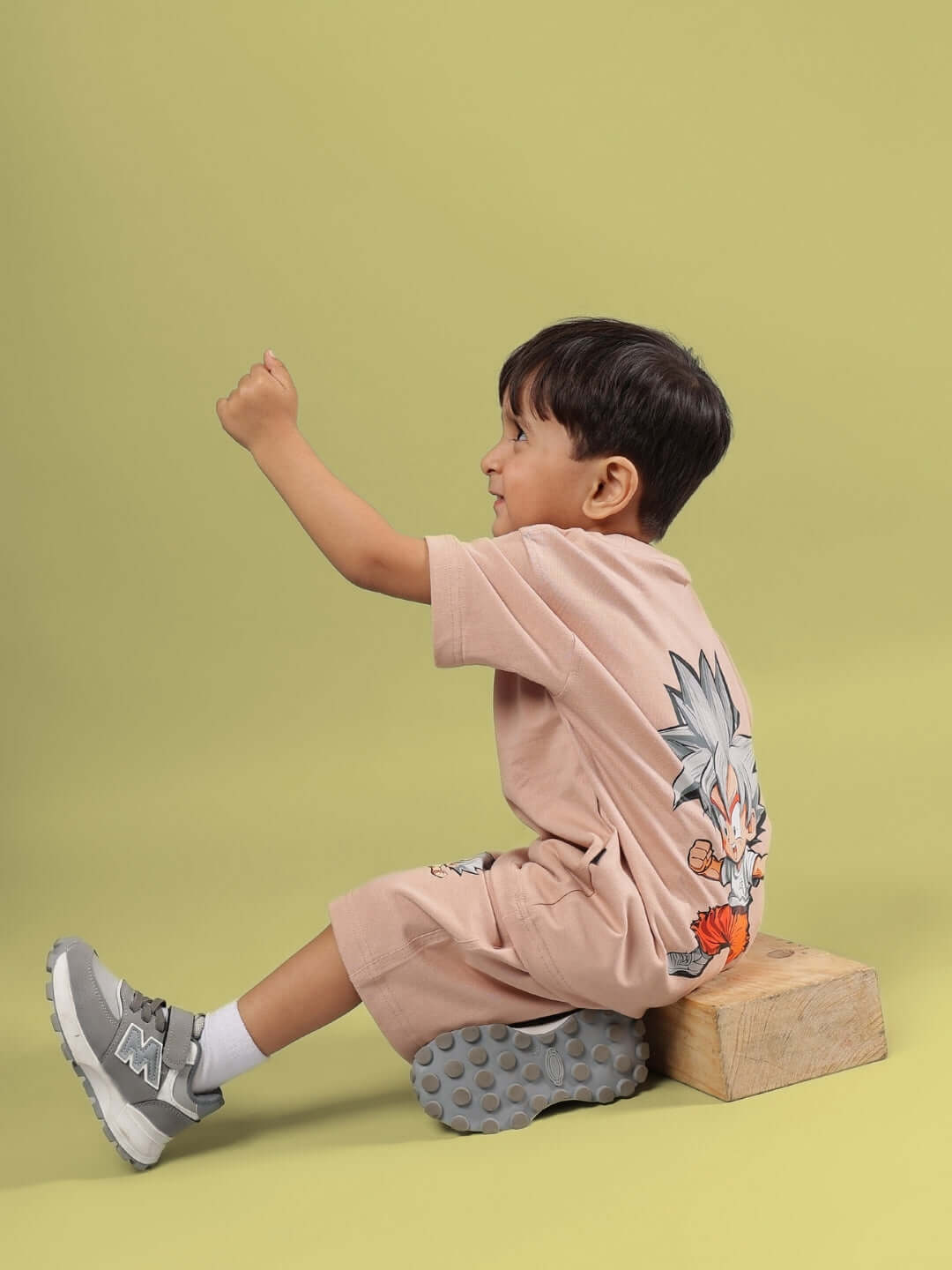Child wearing a beige Gohan t-shirt, sitting on a wooden block, showcasing the back design with character illustration.