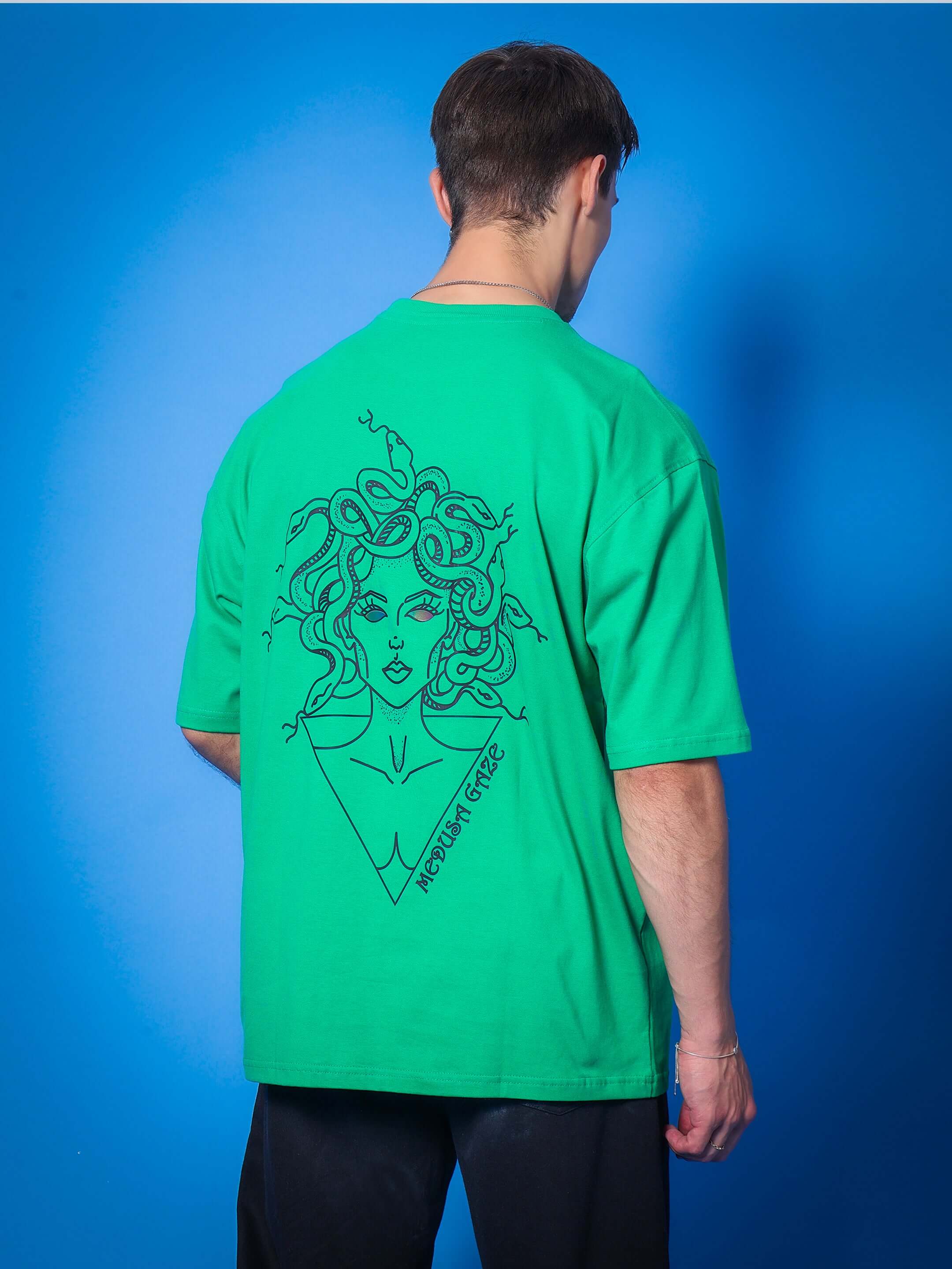 Man wearing MEDUSA GAZE REFLECTOR EYE OVER-SIZED T-SHIRT GREEN with Medusa graphic on back, standing against blue background.