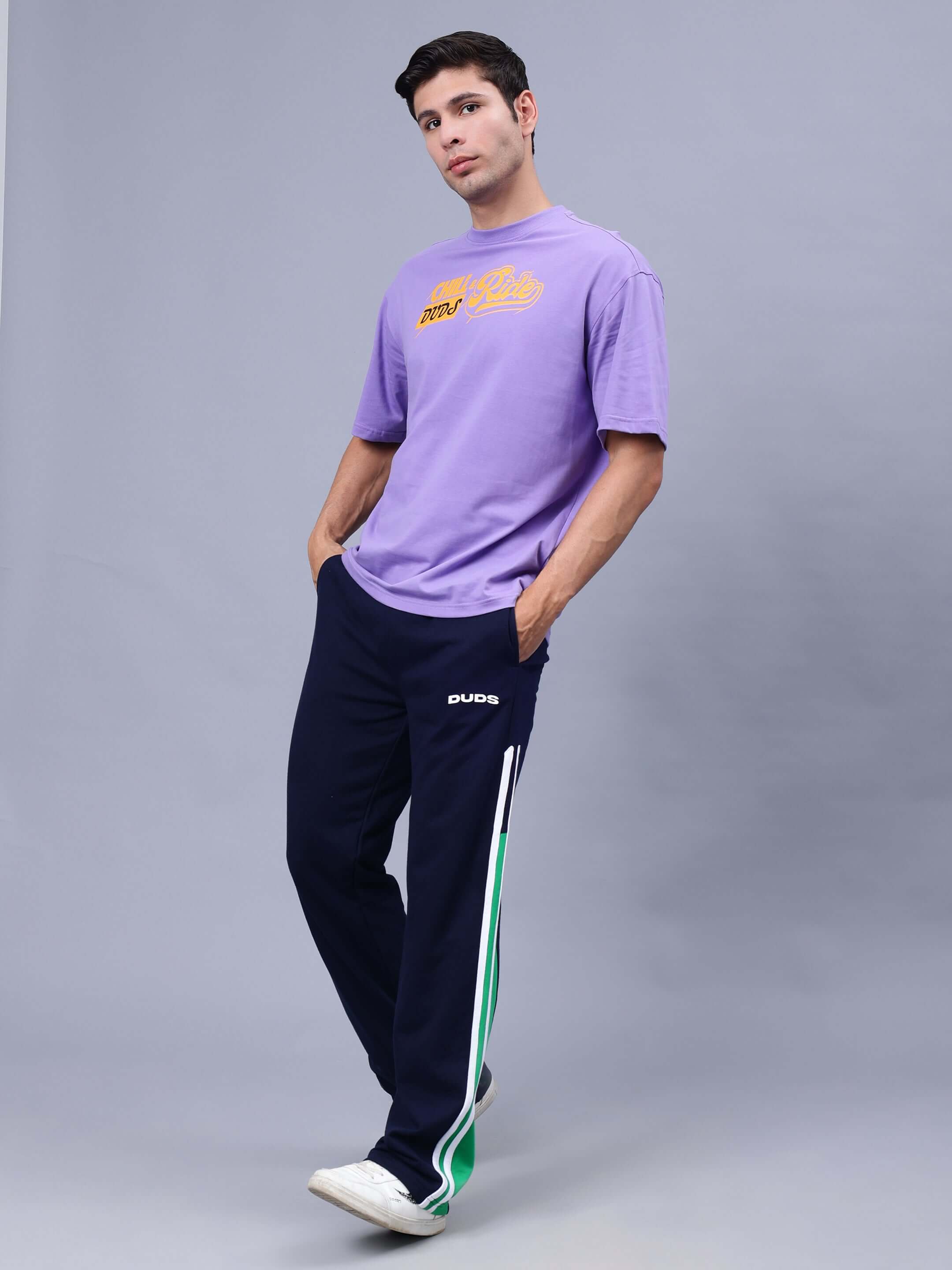 Model wearing DUDS CHILL & RIDE OVER-SIZED T-SHIRT LAVENDER, paired with navy track pants and white sneakers, standing against a gray background.