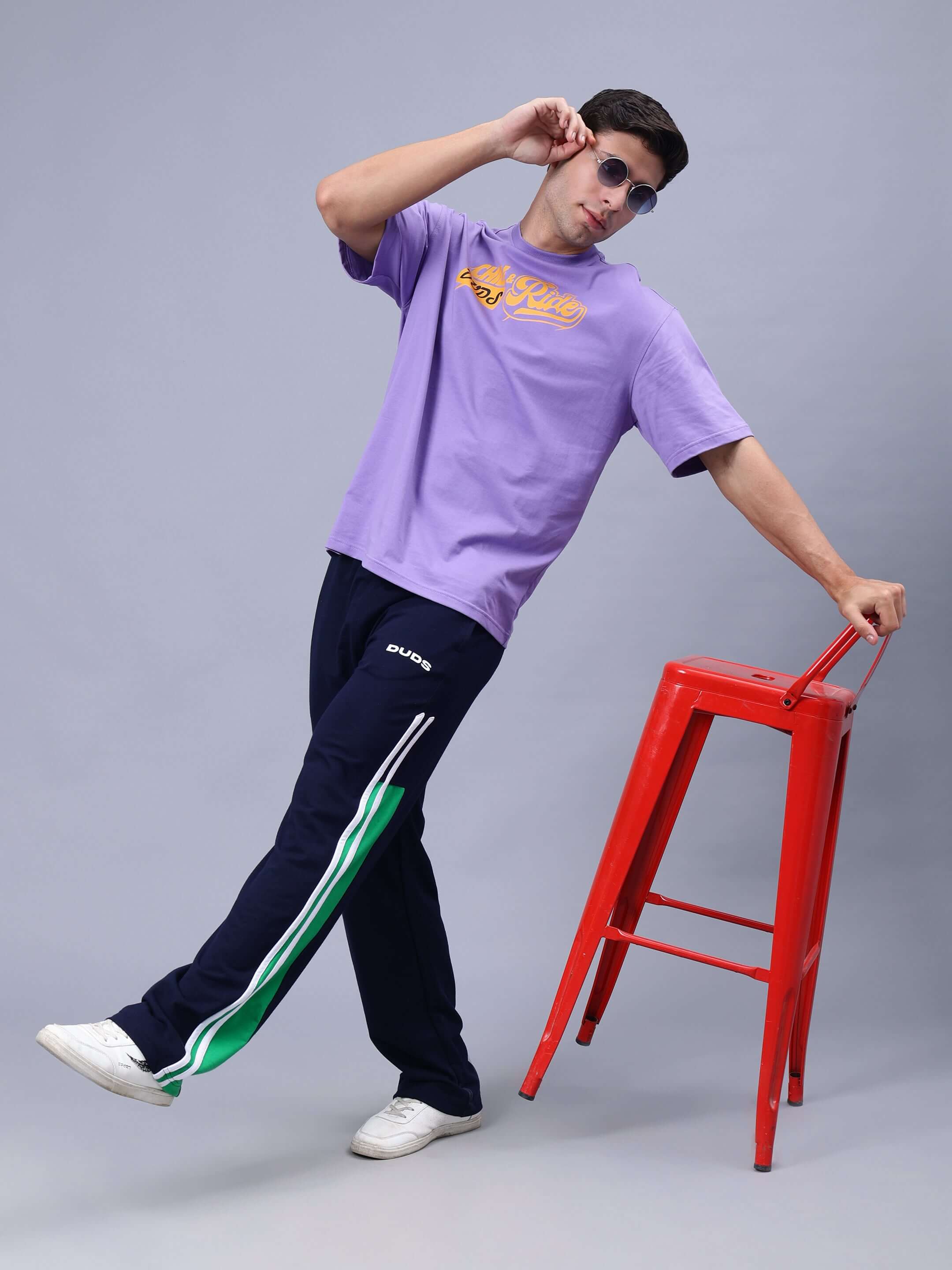 Man modeling DUDS CHILL & RIDE OVER-SIZED T-SHIRT LAVENDER with red stool and sunglasses, emphasizing casual and trendy style.