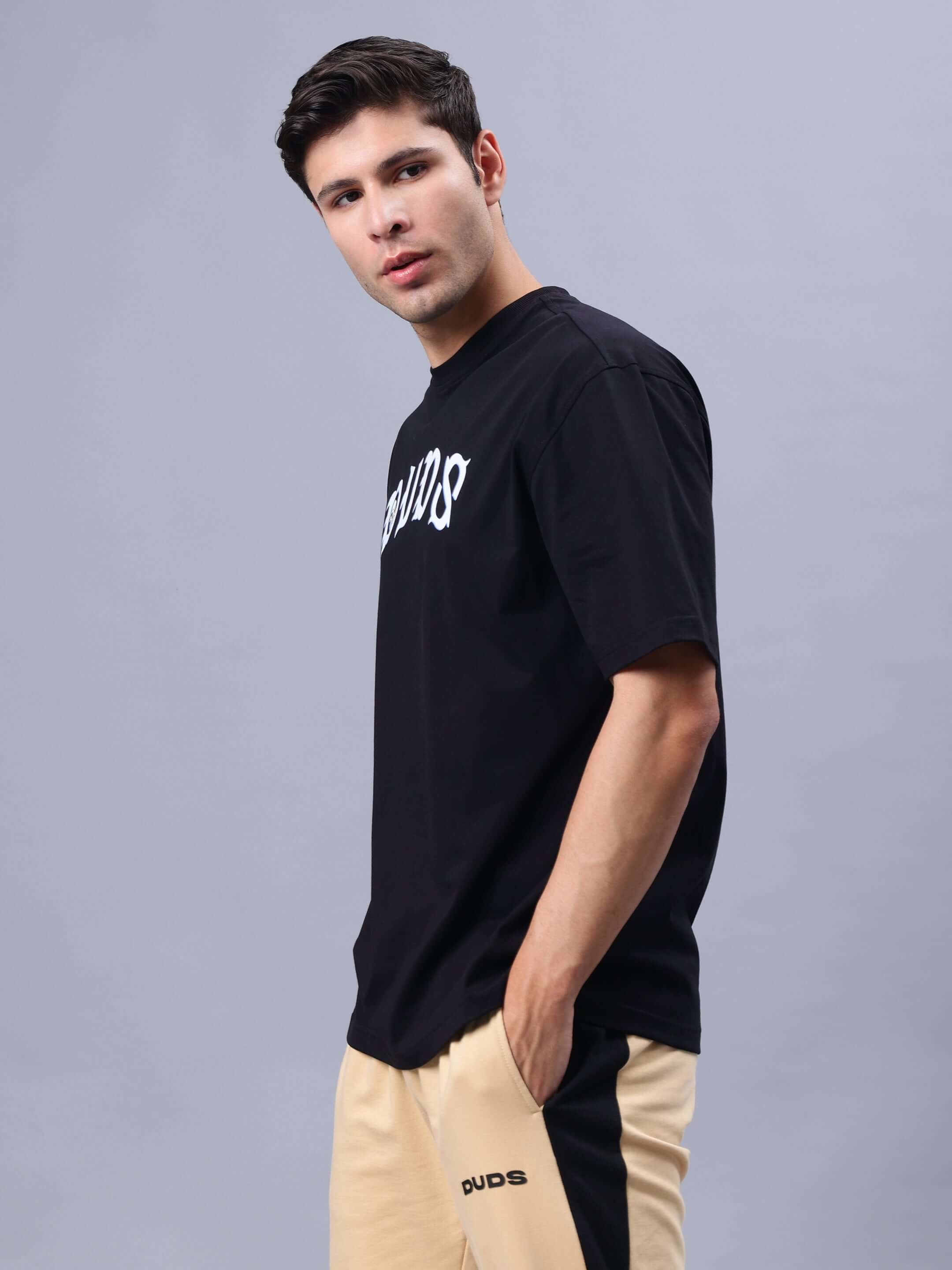 Model wearing REBELS WTF OVER-SIZED T-SHIRT BLACK with hands in pockets, showcasing casual look.
