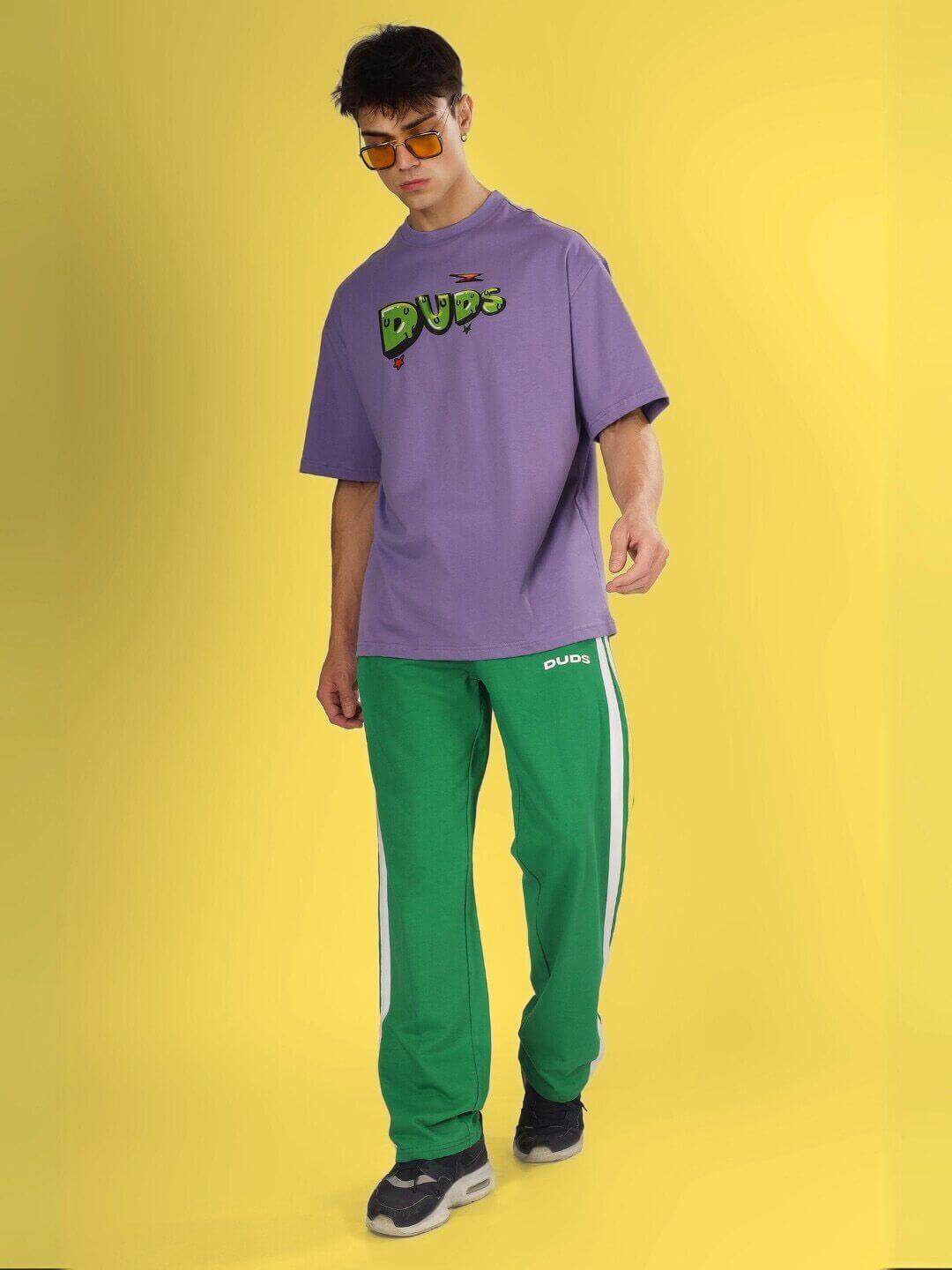 Man wearing ESCAPED TWEETY OVER-SIZED T-SHIRT WISTERIA PURPLE with green pants against yellow background.