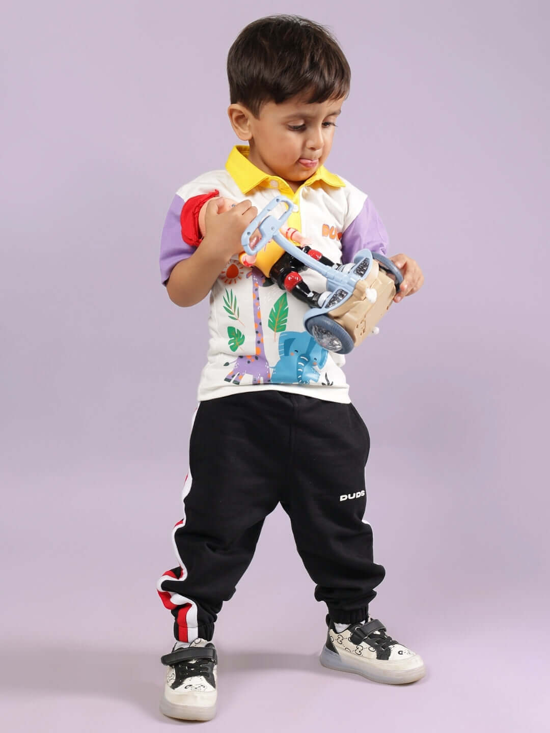 Young boy wearing colorful DUDS outfit, holding a toy drill, standing against a lavender background.