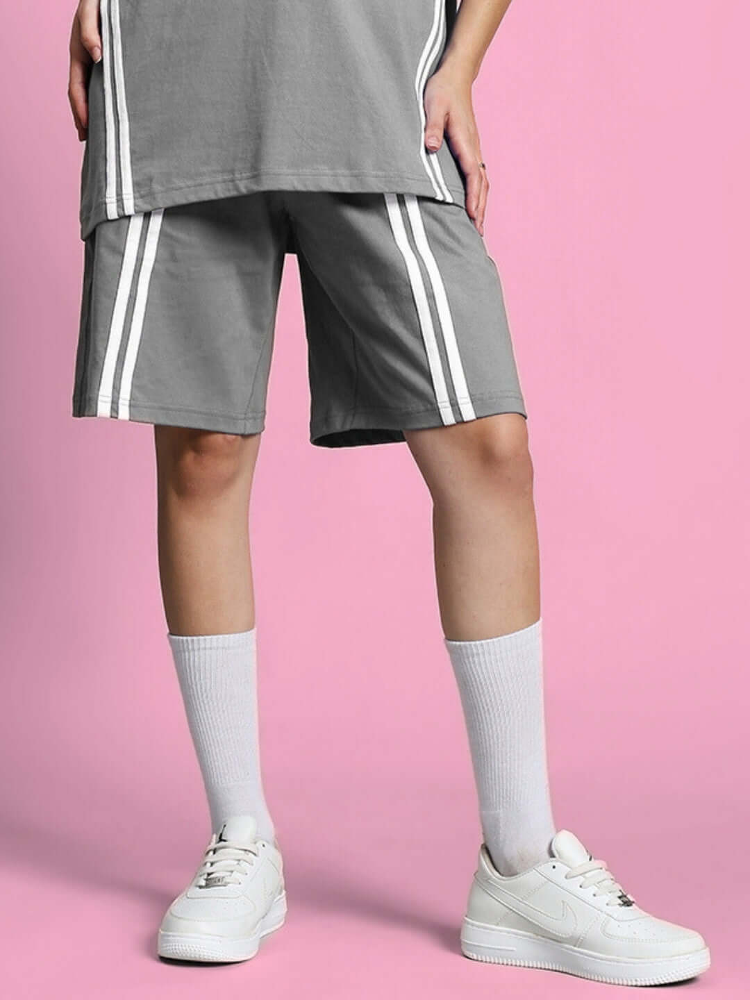 Women's grey regular fit shorts with white stripes, styled with white sneakers and socks against a pink background.