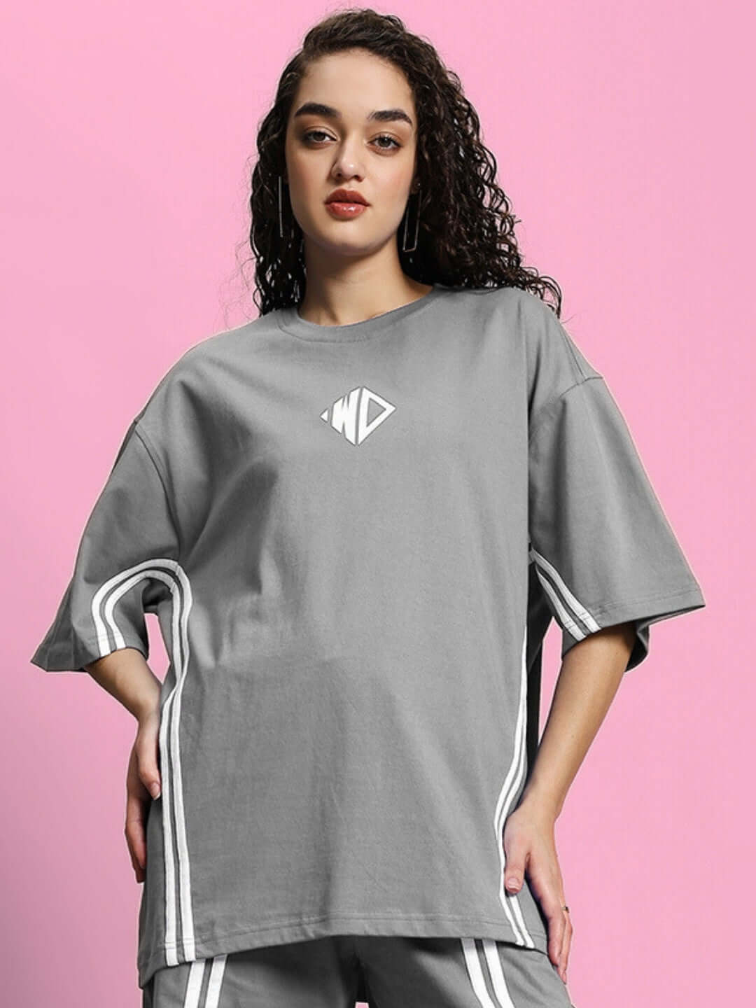 Woman wearing a grey oversized T-shirt made of premium cotton fabric, featuring a relaxed fit and white stripe details, against a pink background.