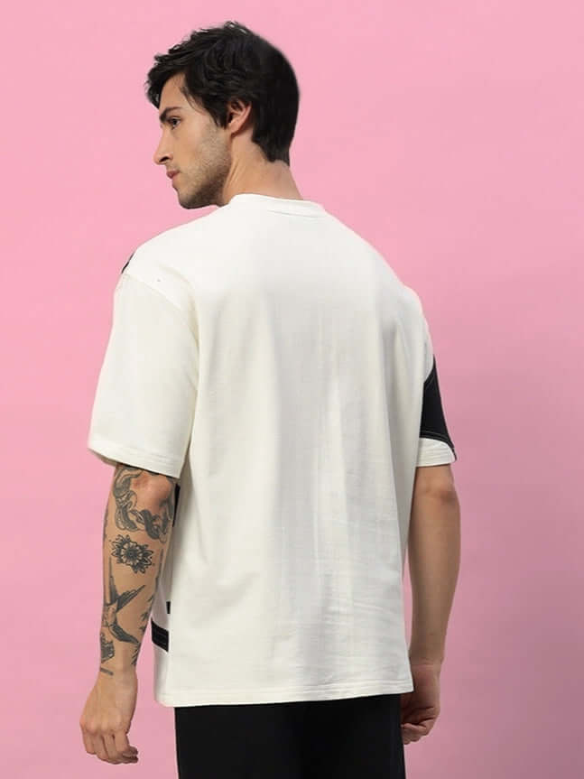 Back view of a man wearing a DAVID Designer Oversized T-shirt in off white-black with tattooed arm, against a pink background.
