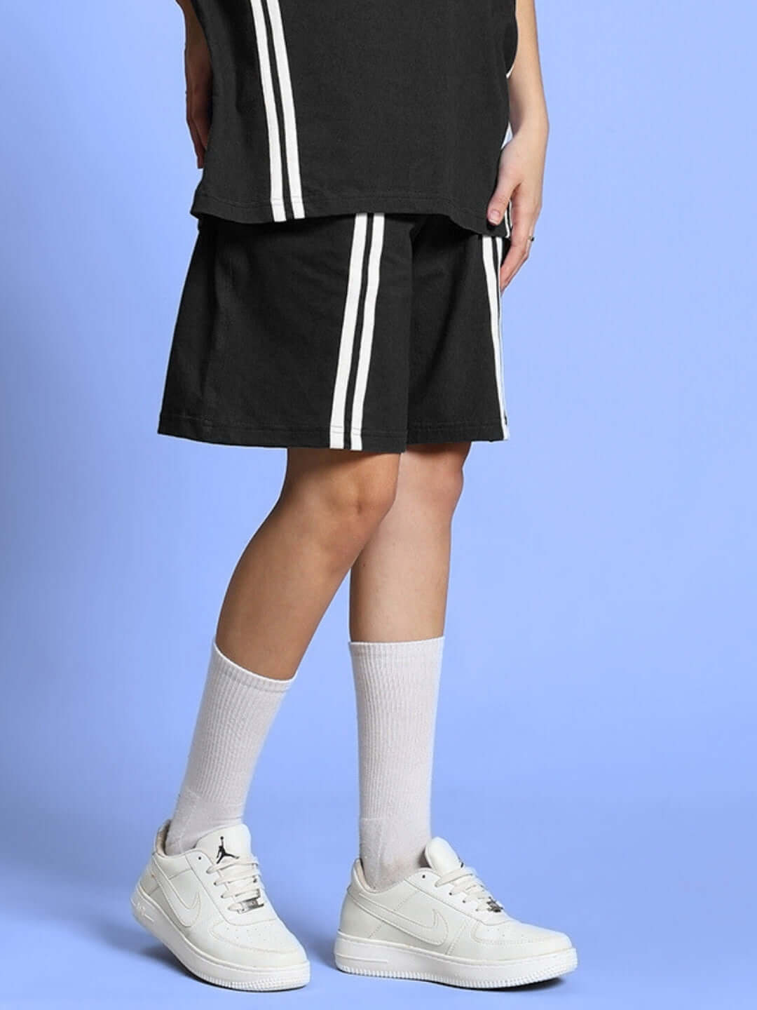 Women's Outlook Regular Fit Black Shorts with White Stripes, Ideal for Custom Fit, Paired with Sneakers, Shot Against Blue Background.