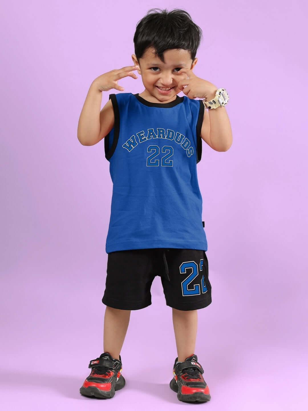 Child wearing Amiga black and blue shorts set from DUDS, smiling with playful pose, on a light purple background.