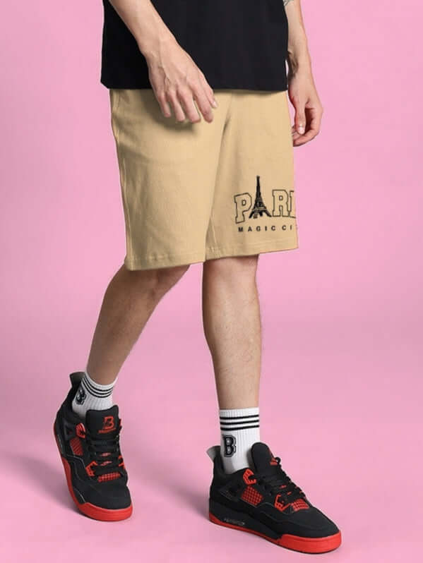 Person wearing beige MAGIC CITY SUMMER SHORTS with Paris emblem and black sneakers, standing against a pink background.