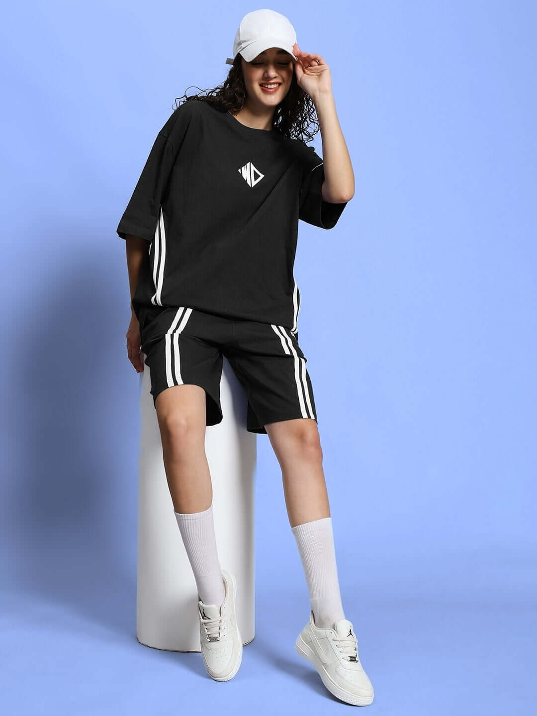 Woman wearing black regular fit shorts and matching top, styled with white socks and sneakers, posing cheerfully against a blue background.