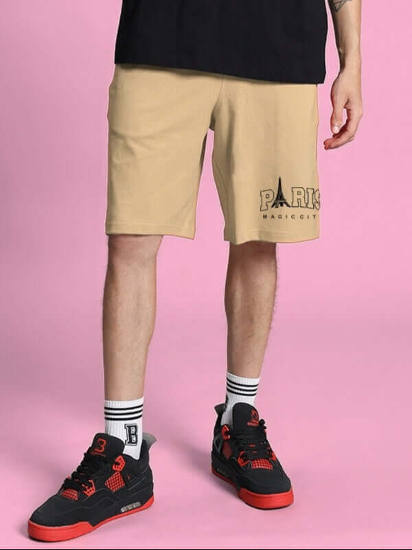 Model wearing beige Magic City Summer Shorts with Paris logo, paired with black and red sneakers against a pink background.