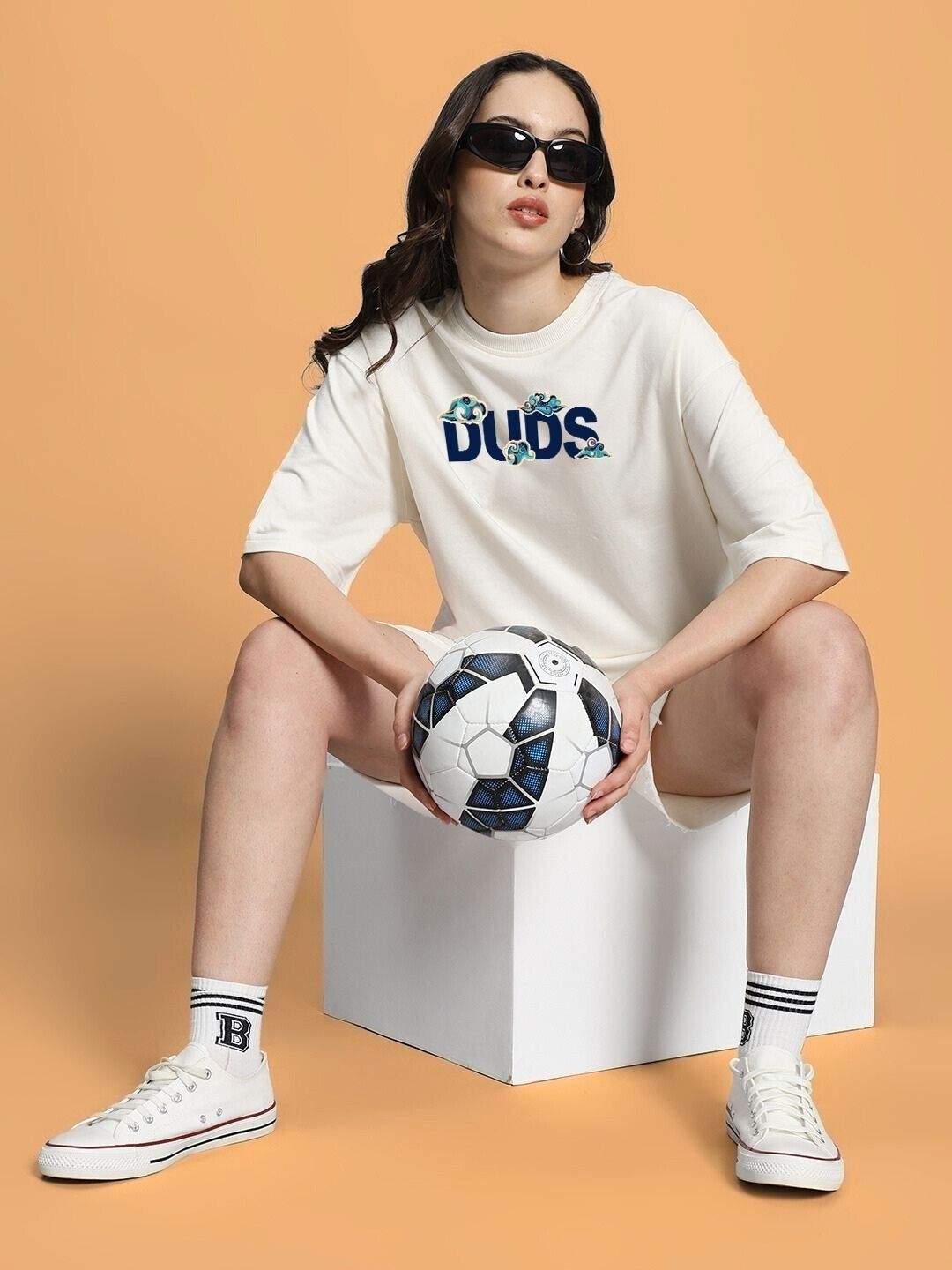 Woman in casual outfit sitting on a box holding a soccer ball, wearing sporty shoes and sunglasses against an orange background.