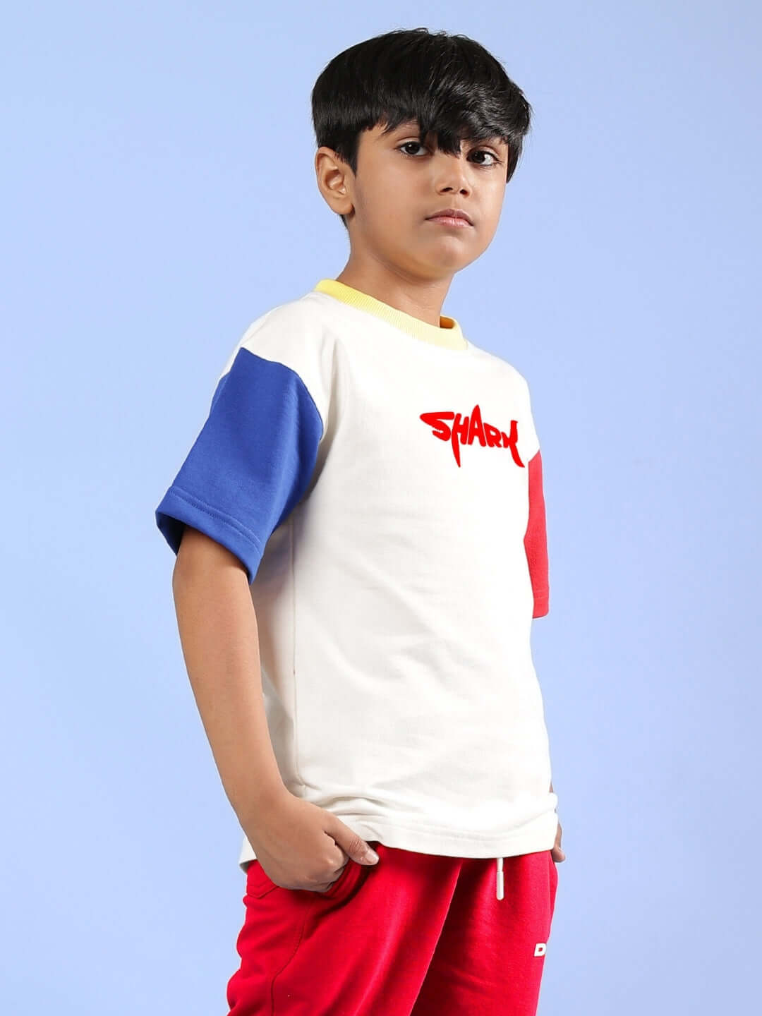Boy wearing off-white shark fish printed oversized T-shirt with blue and red sleeves and red shorts, against a blue background.